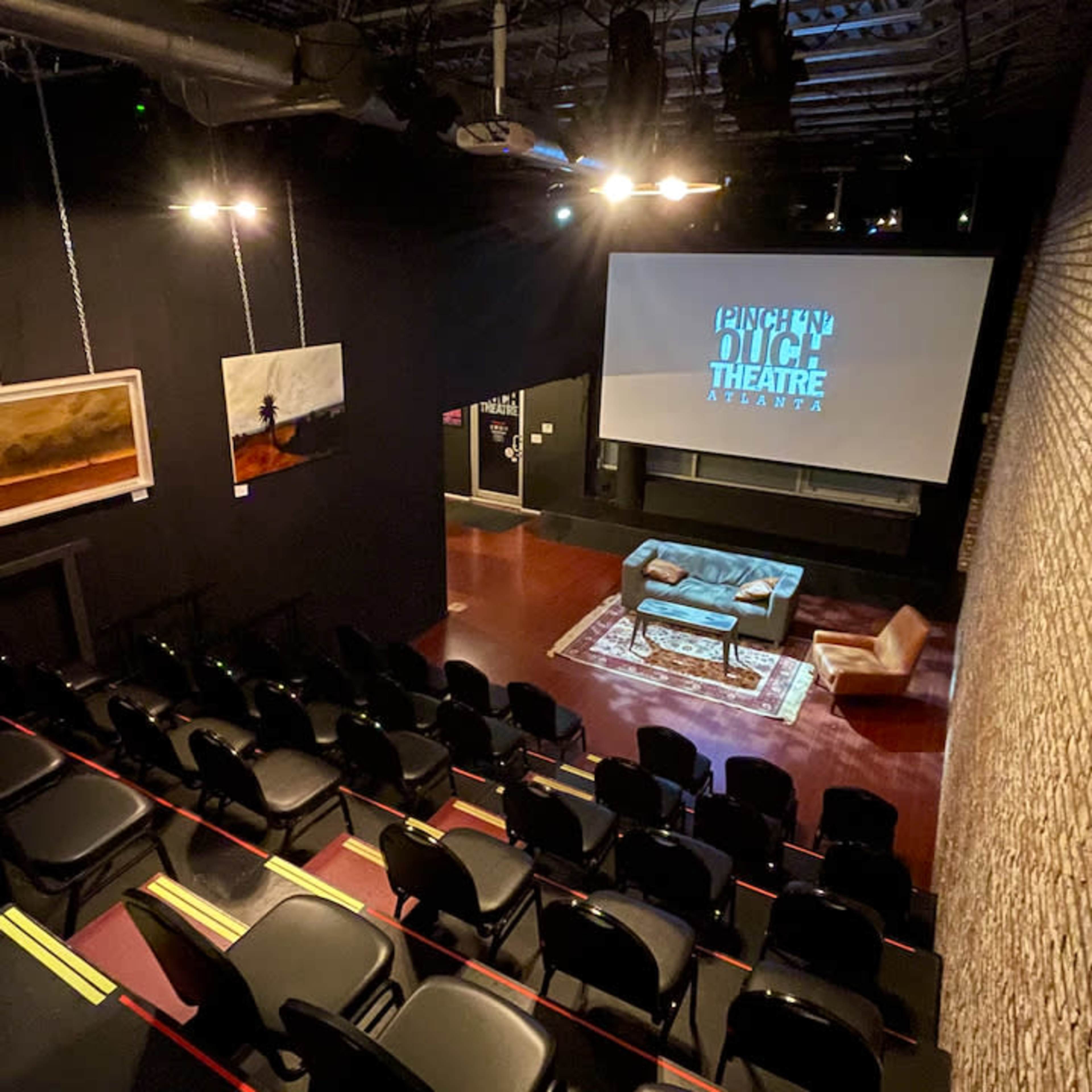 An interior view of a small theater featuring rows of black seating, a large screen displaying the theater's name, and a stage with a sofa and artwork.