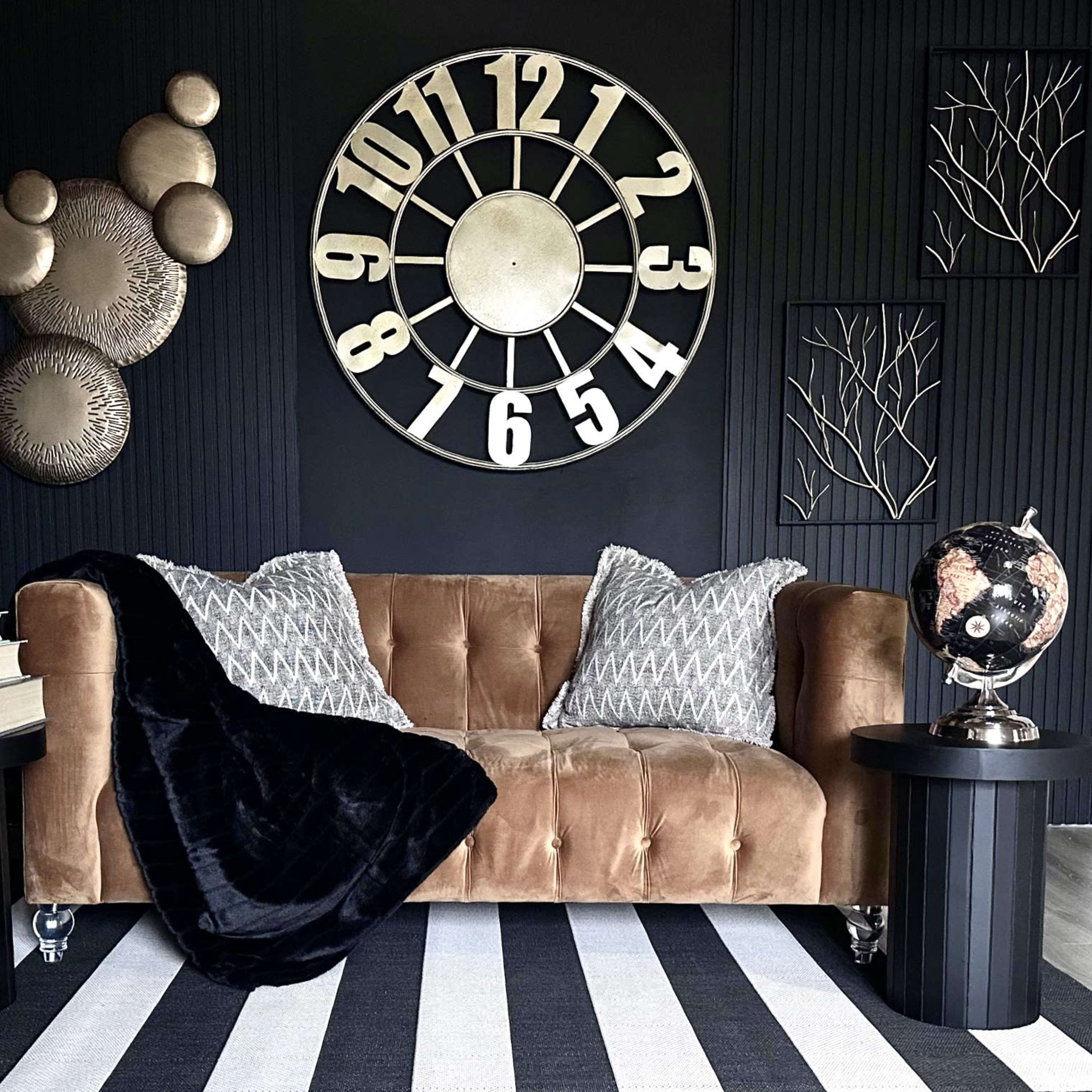 A brown couch with decorative pillows sits between two black side tables, featuring a large wall clock and metallic art pieces against a dark background.