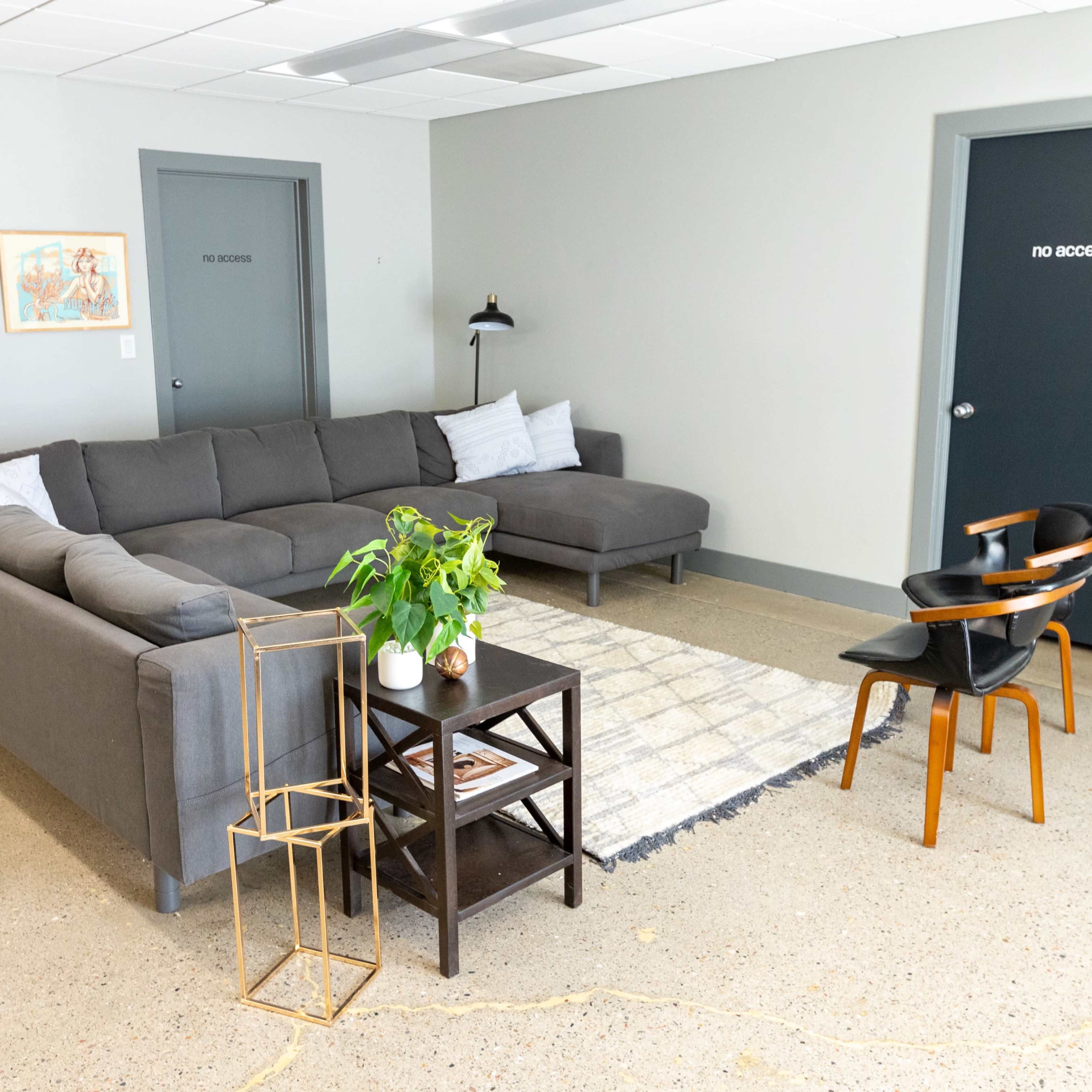 A modern lounge area with a gray sectional sofa, a small black side table, a plant, and two chairs, set against a minimalistic backdrop with a closed door labeled "NO ACCESS."