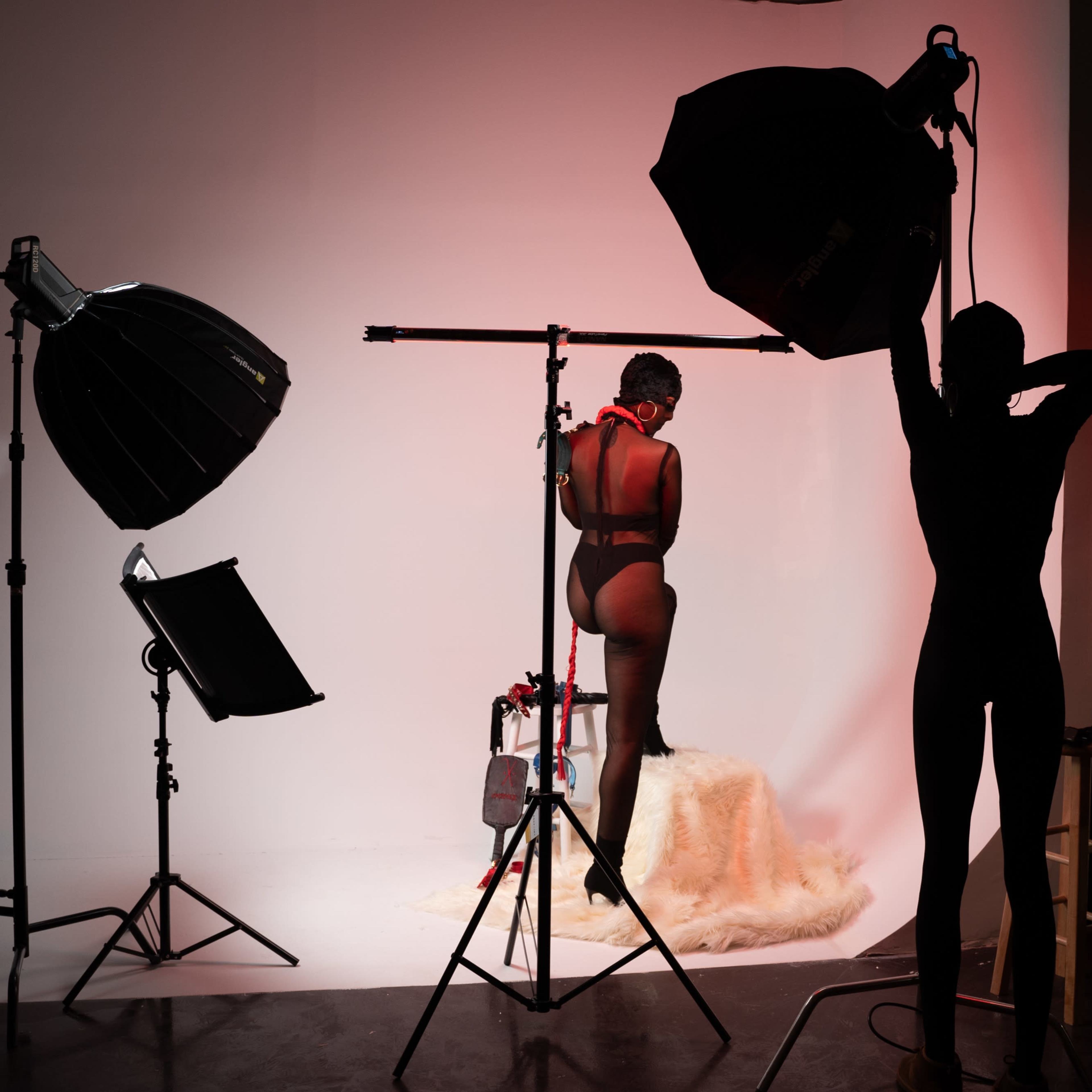 A woman poses on a stool in a photography studio, surrounded by lighting equipment against a plain backdrop.
