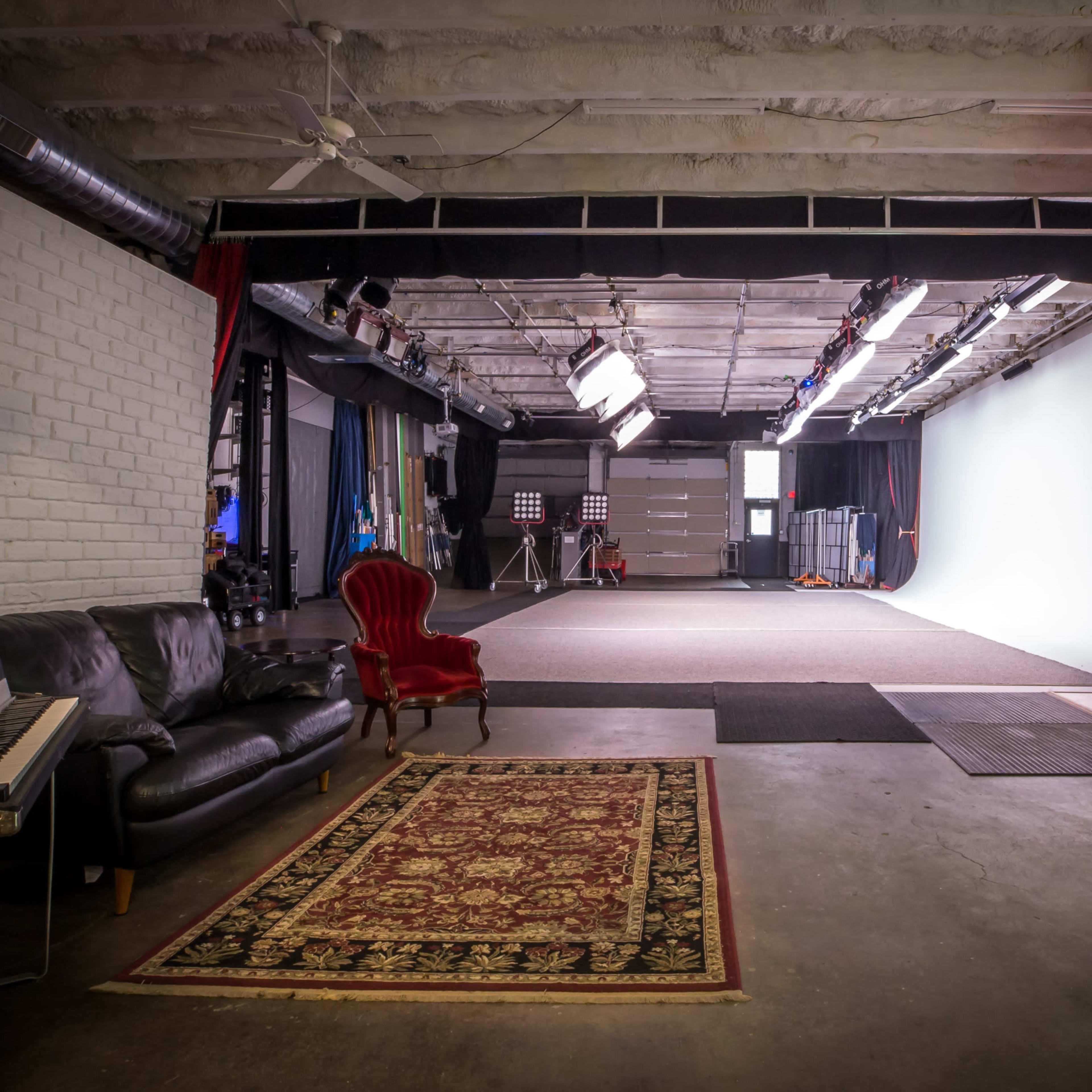 A film studio with a seating area featuring a black sofa and a red chair, leading to a large empty space with a white backdrop and studio lighting.