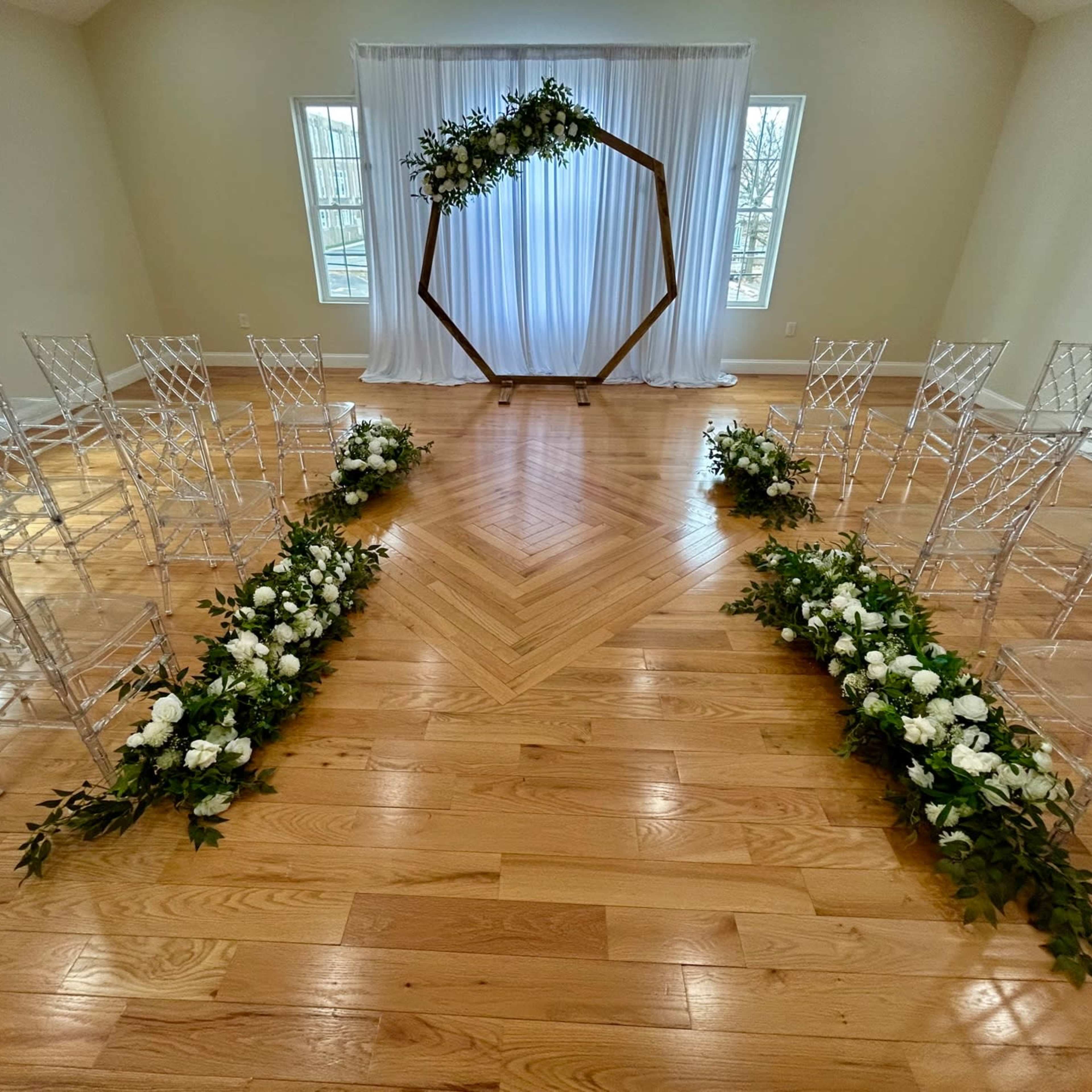 A wedding ceremony setup with transparent chairs arranged in two rows facing an ornate wooden arch decorated with greenery and white flowers, in front of a backdrop of flowing white fabric.