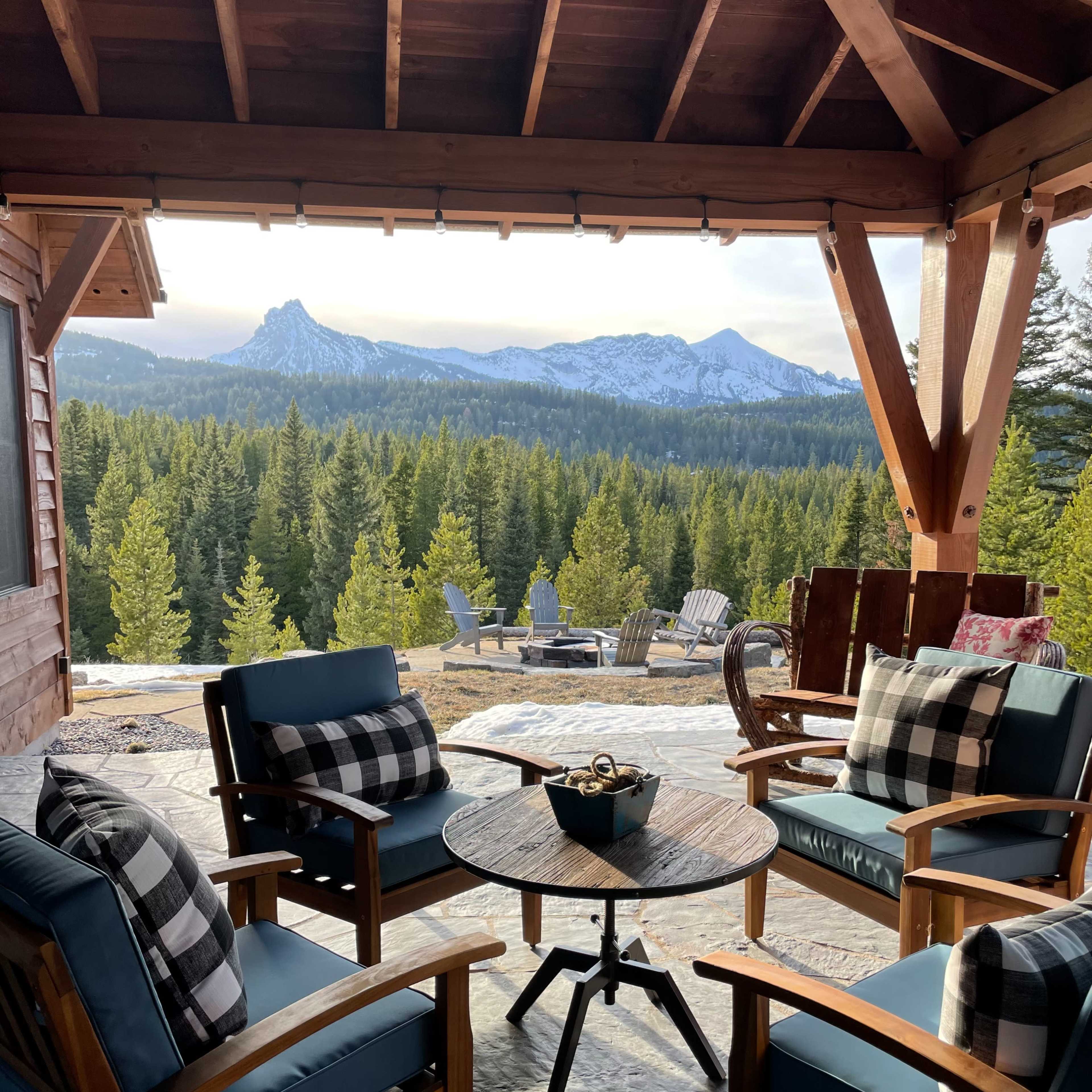 A cozy outdoor seating area features wooden chairs and a round table, overlooking a forested landscape and snowy mountains in the distance.