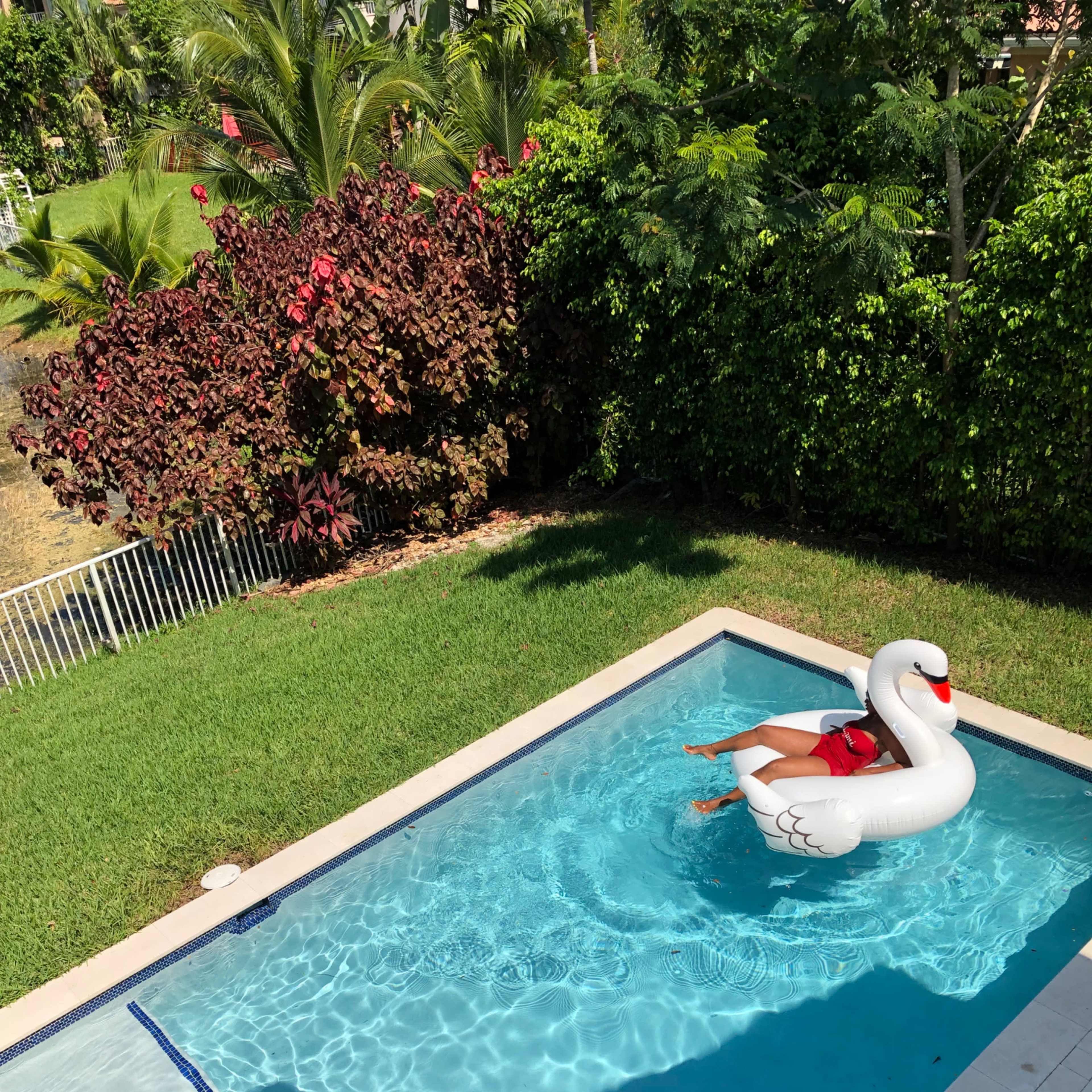 A person wearing a red swimsuit relaxes on a large white swan float in a swimming pool surrounded by greenery.