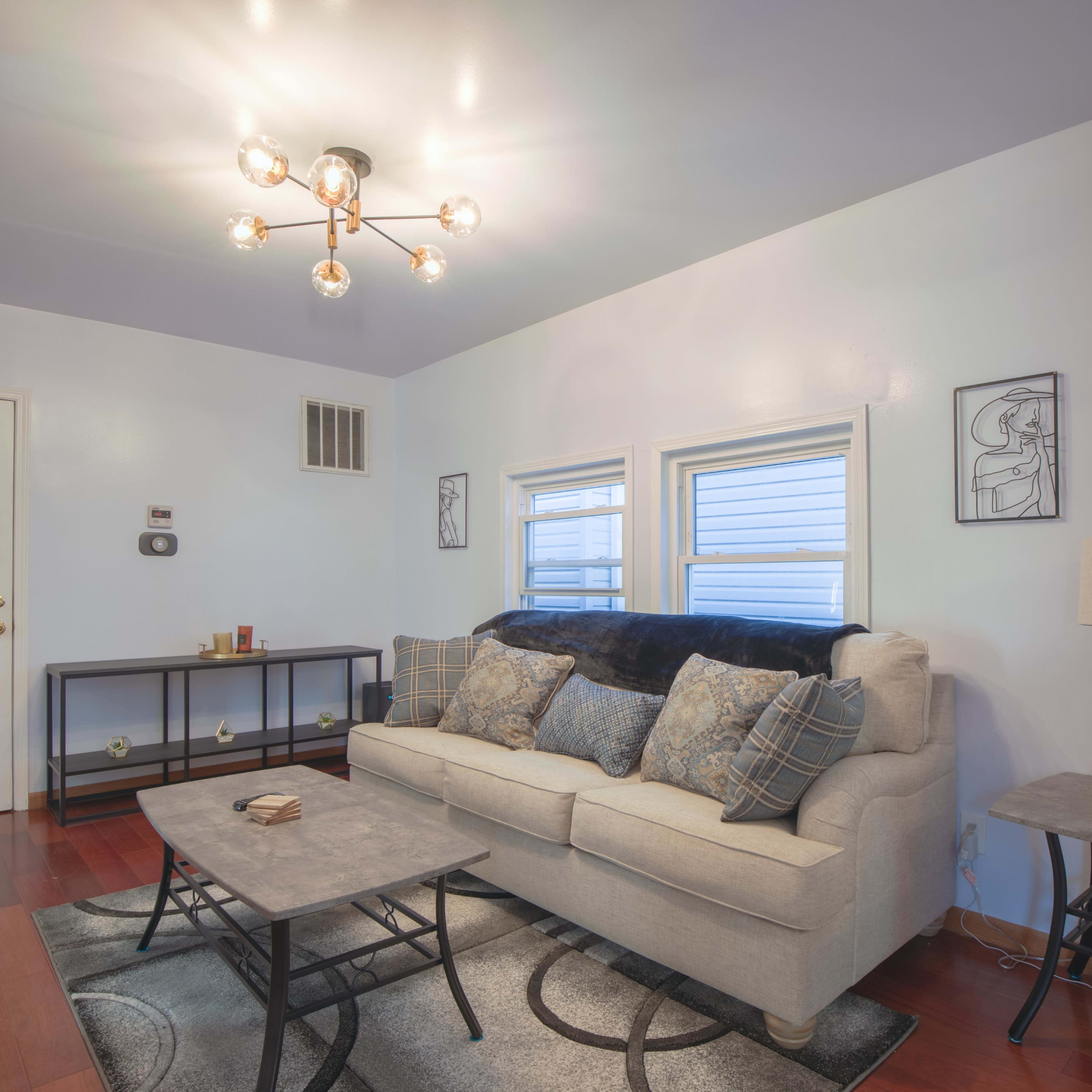 A modern living room features a light-colored sofa with patterned pillows, a round coffee table, and a lamp beside a decorative globe, all set against a wall with two windows and a door.
