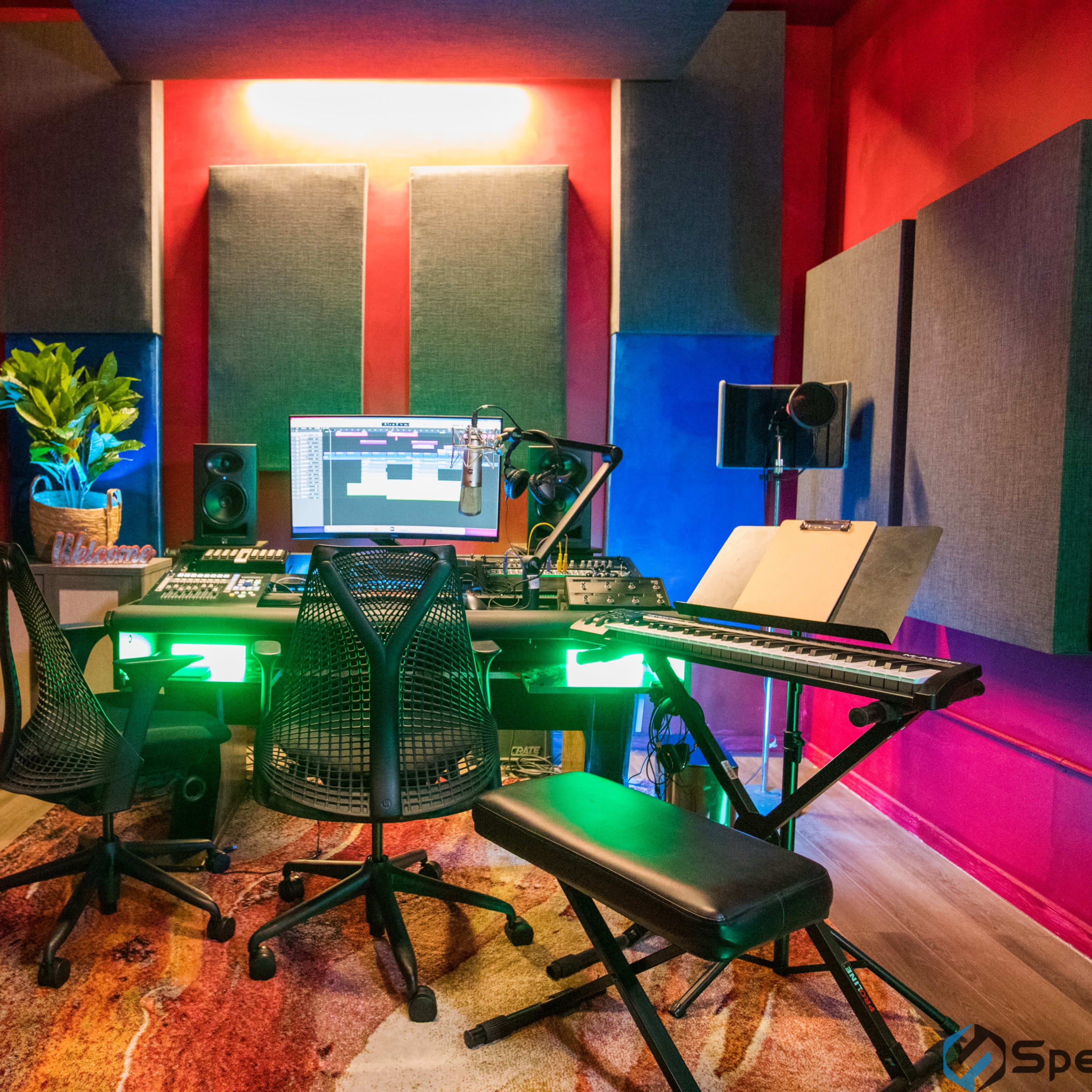 A music production studio features a central desk with a computer setup, surrounded by soundproofing panels, instruments on the walls, and a colorful, illuminated atmosphere.