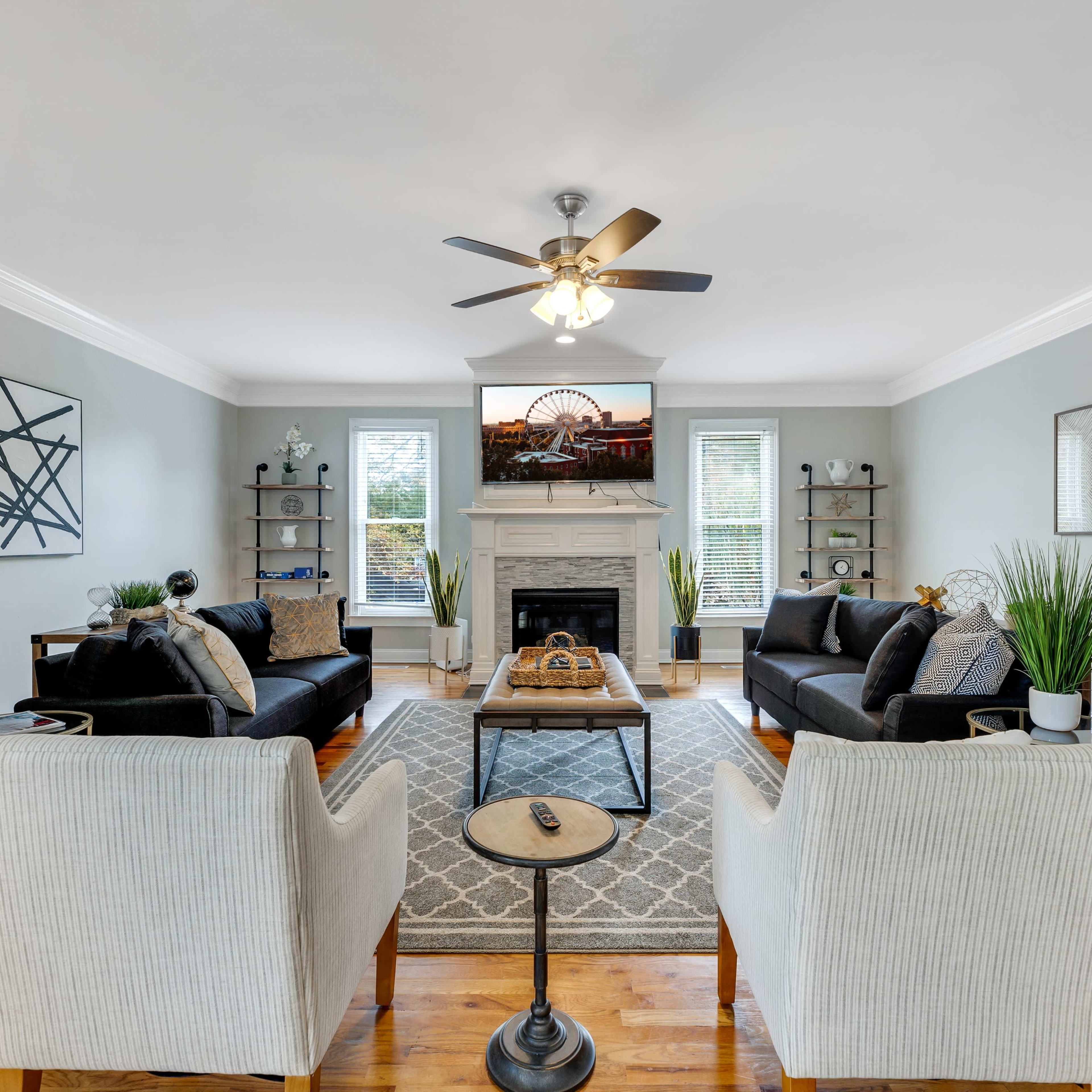 A spacious living room features two black sofas, two light-colored armchairs, a coffee table, and a fireplace, with decorative artwork on the walls and plants scattered throughout.
