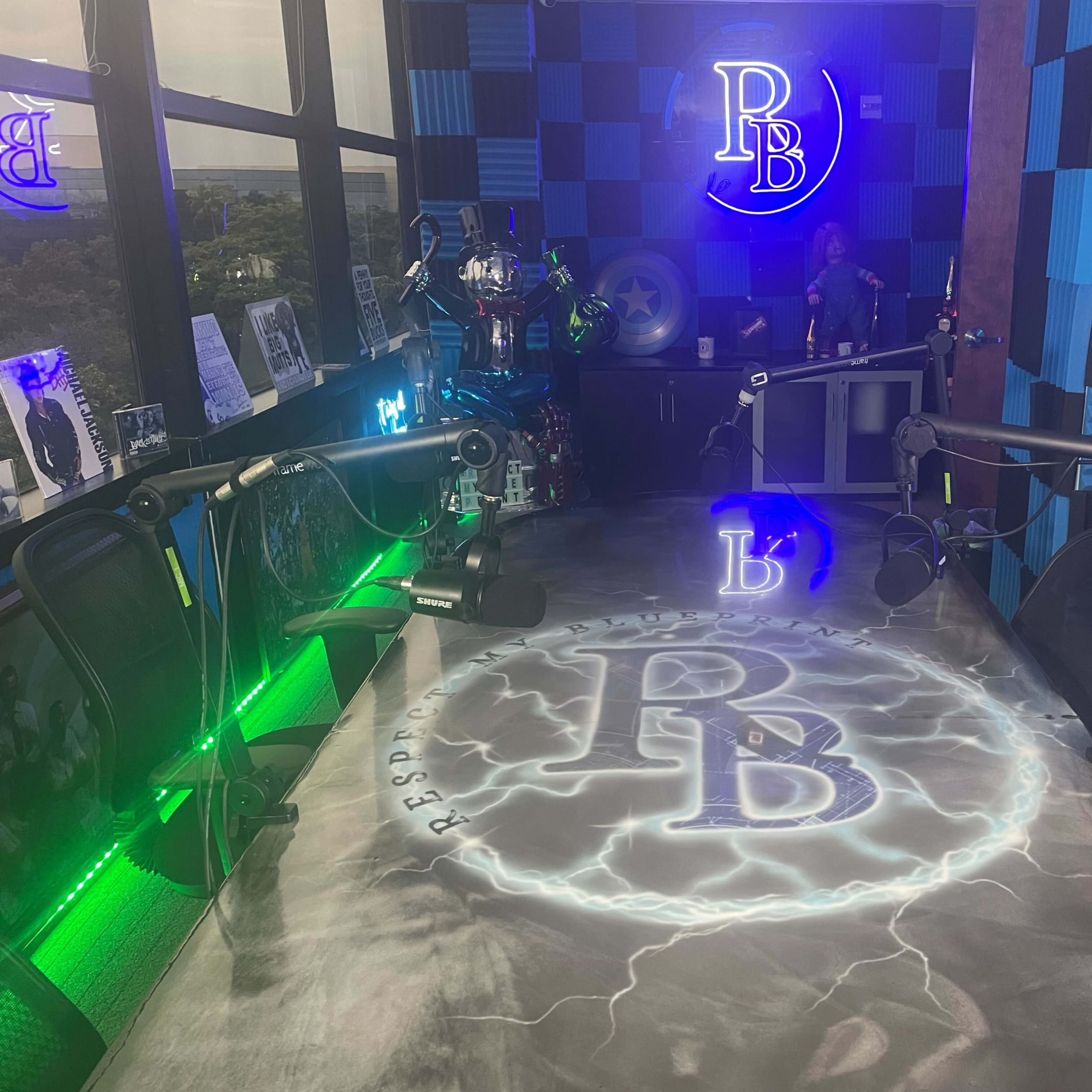 A brightly lit recording studio with a large table featuring the letters "PB" in a lightning design, surrounded by microphone stands and colorful acoustic paneling.