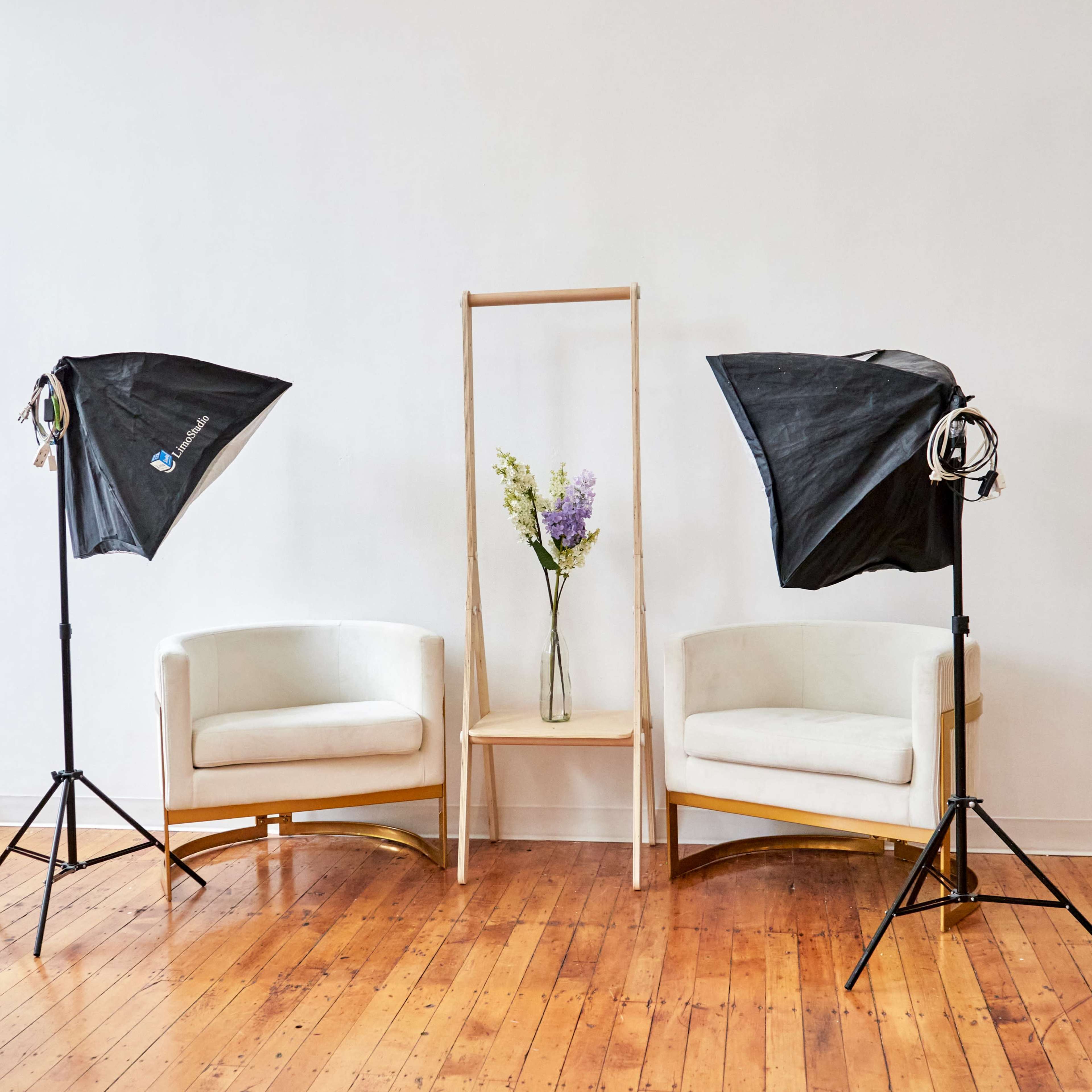 Two white chairs positioned on a wooden floor, with a simple wooden stand holding a vase of flowers between two studio lighting setups.