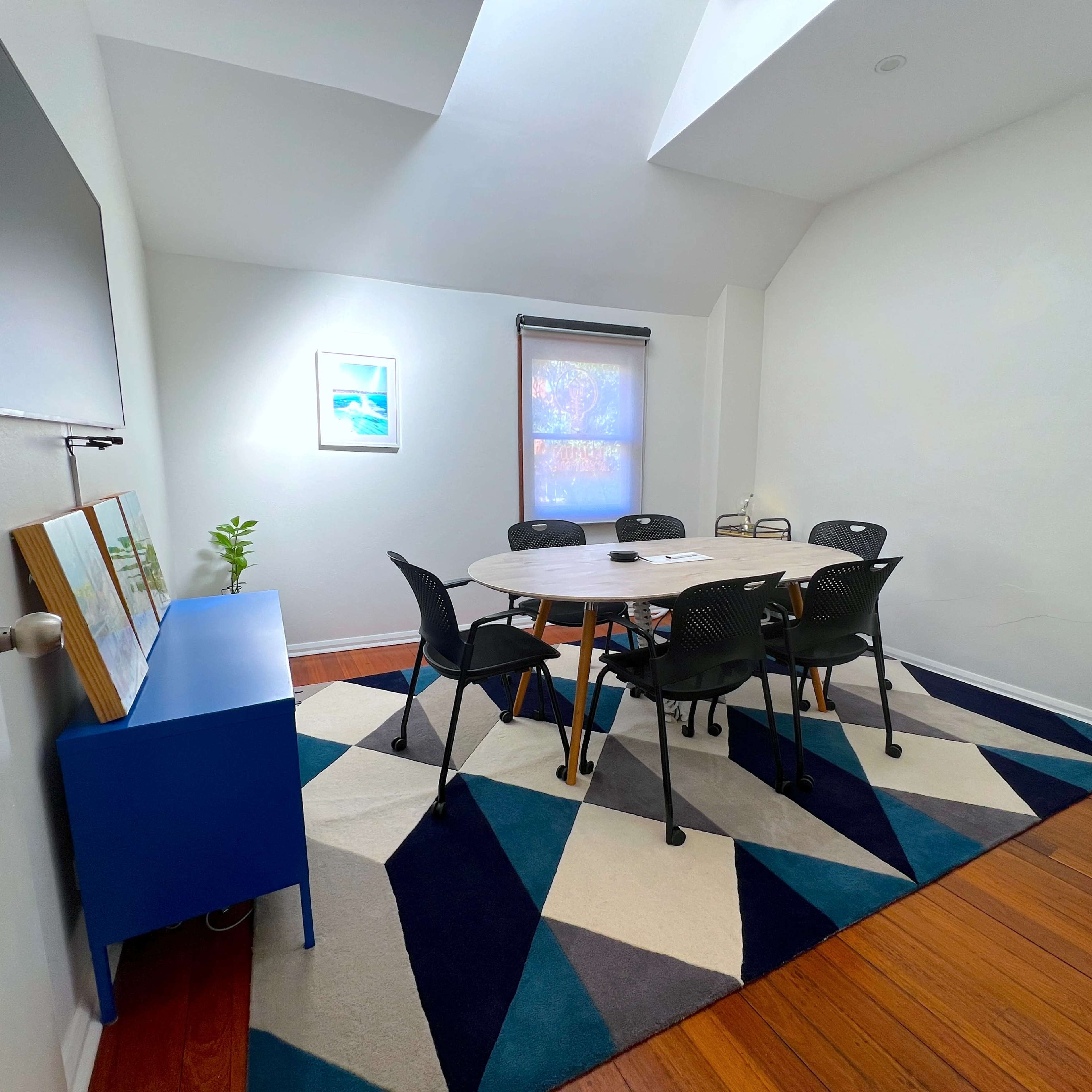 A conference room features a round table surrounded by black chairs on a patterned rug, with a television mounted on one wall and a plant on a blue sideboard.