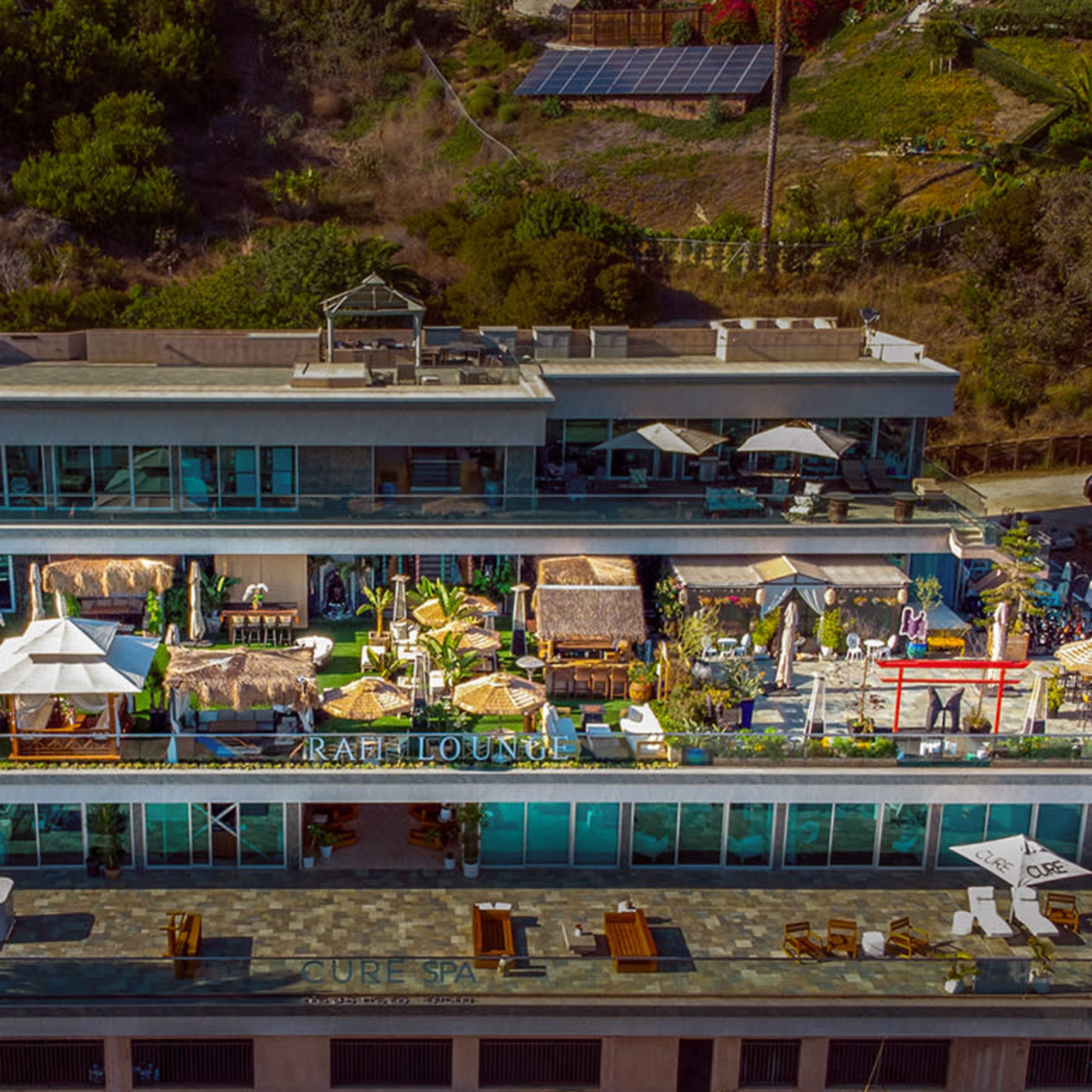 A multi-level building with a rooftop terrace featuring various lounge areas, seating, and decorative structures surrounded by greenery.