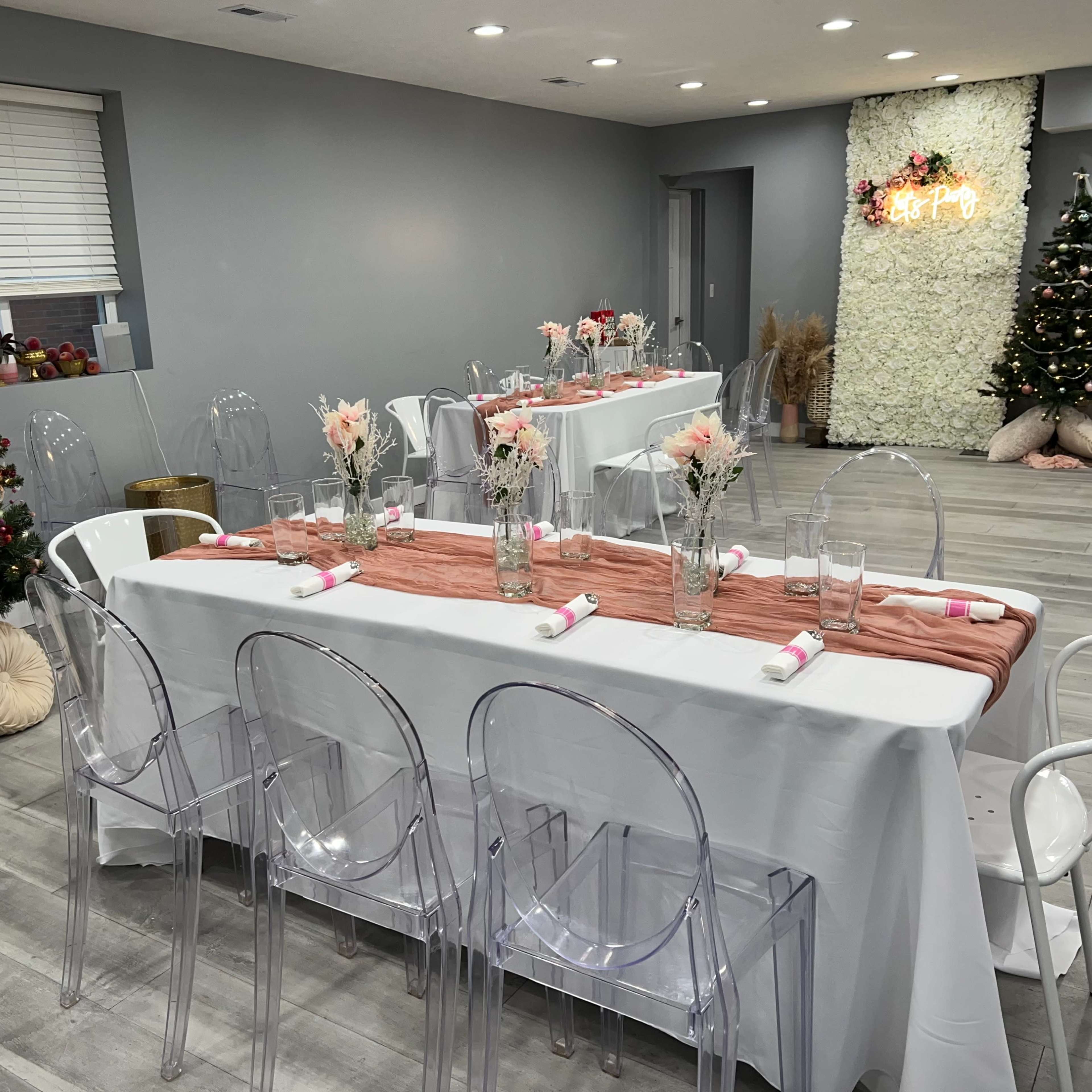 A dining area is set up with clear chairs, white tables covered in linens, and decorated with floral centerpieces alongside two Christmas trees.