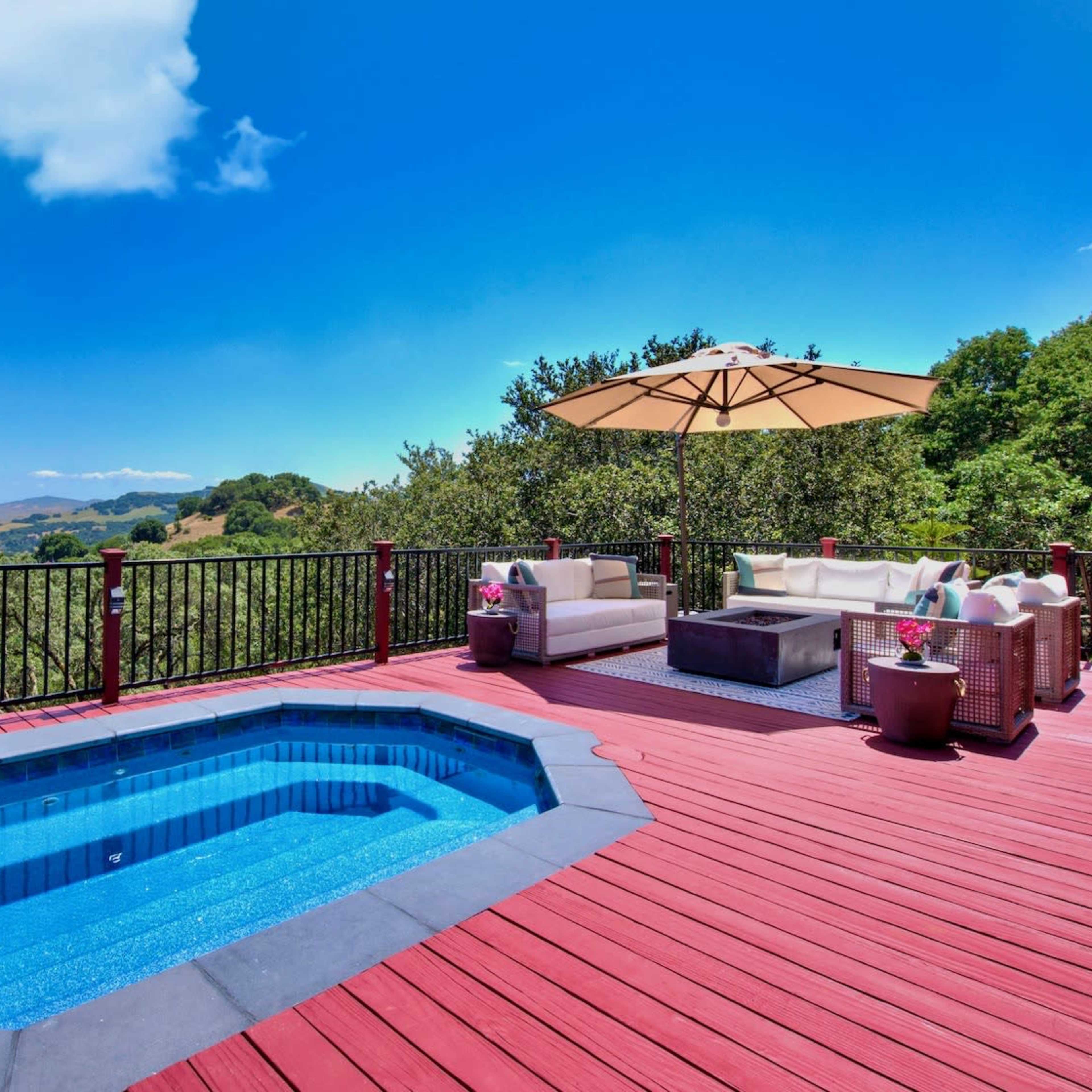 A hot tub is situated on a wooden deck with outdoor seating and an umbrella, overlooking a scenic landscape.