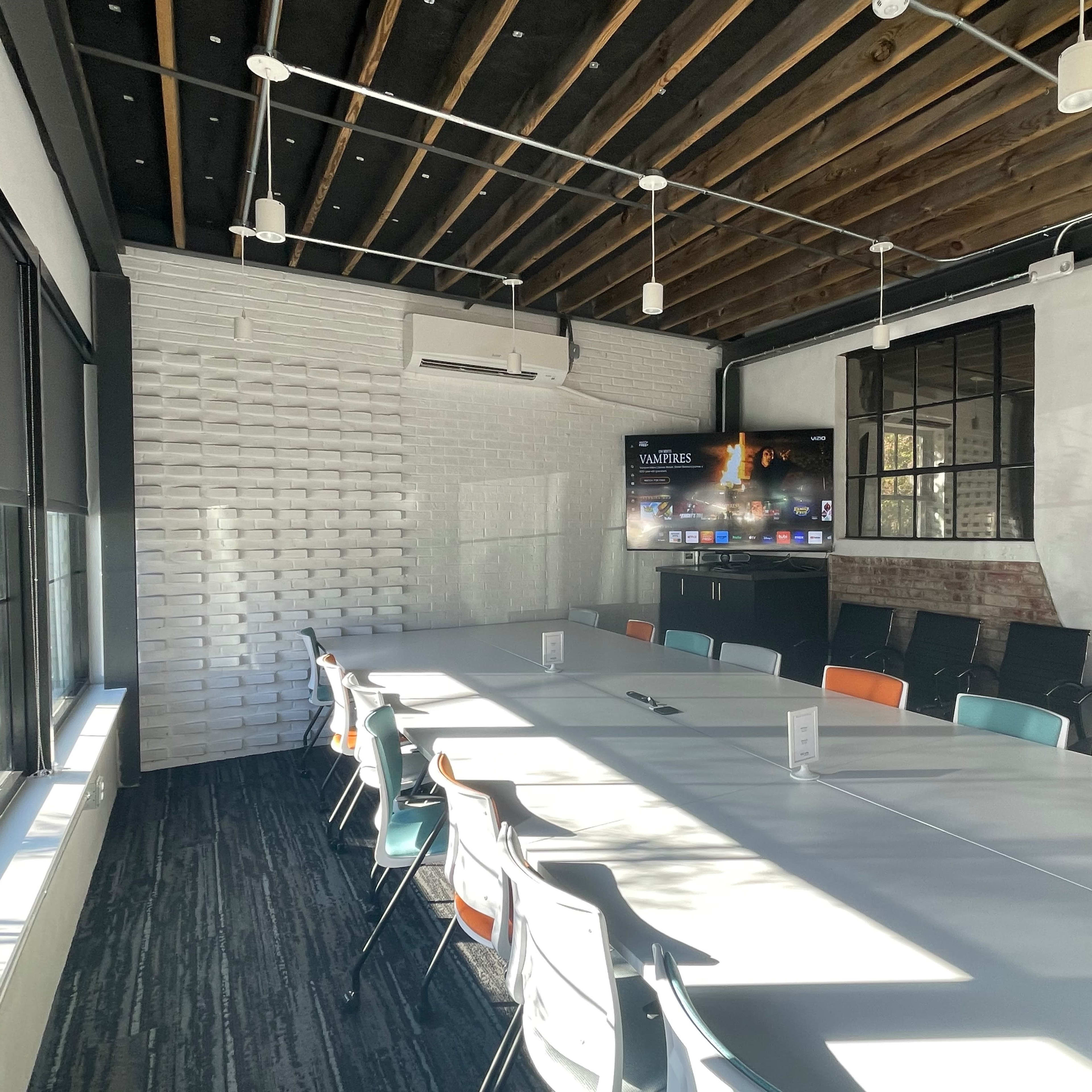 A modern conference room with a long table surrounded by chairs, featuring a large screen on one wall and windows allowing natural light to enter.