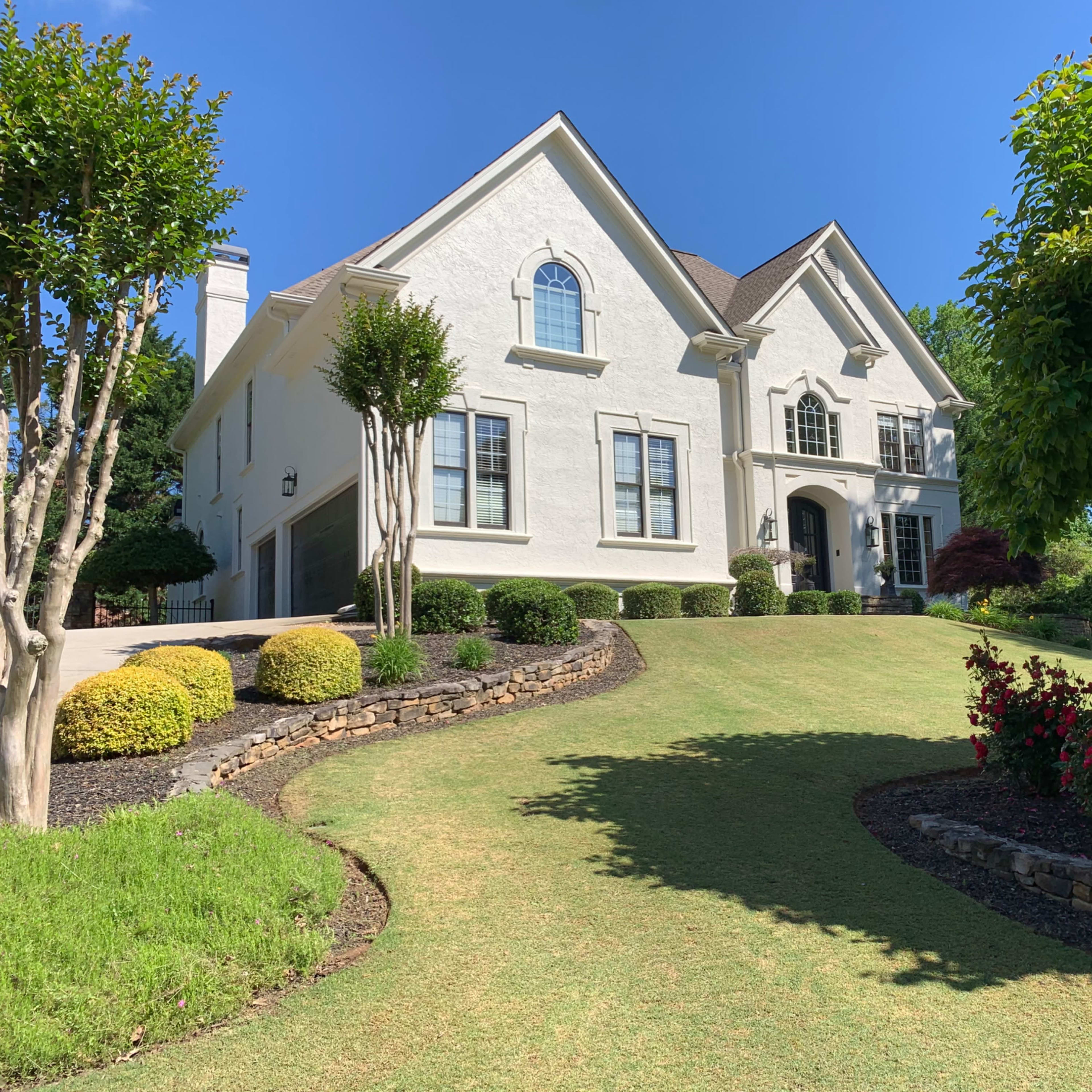 A large, white two-story house with a well-manicured lawn and landscaped flower beds is set against a clear blue sky.