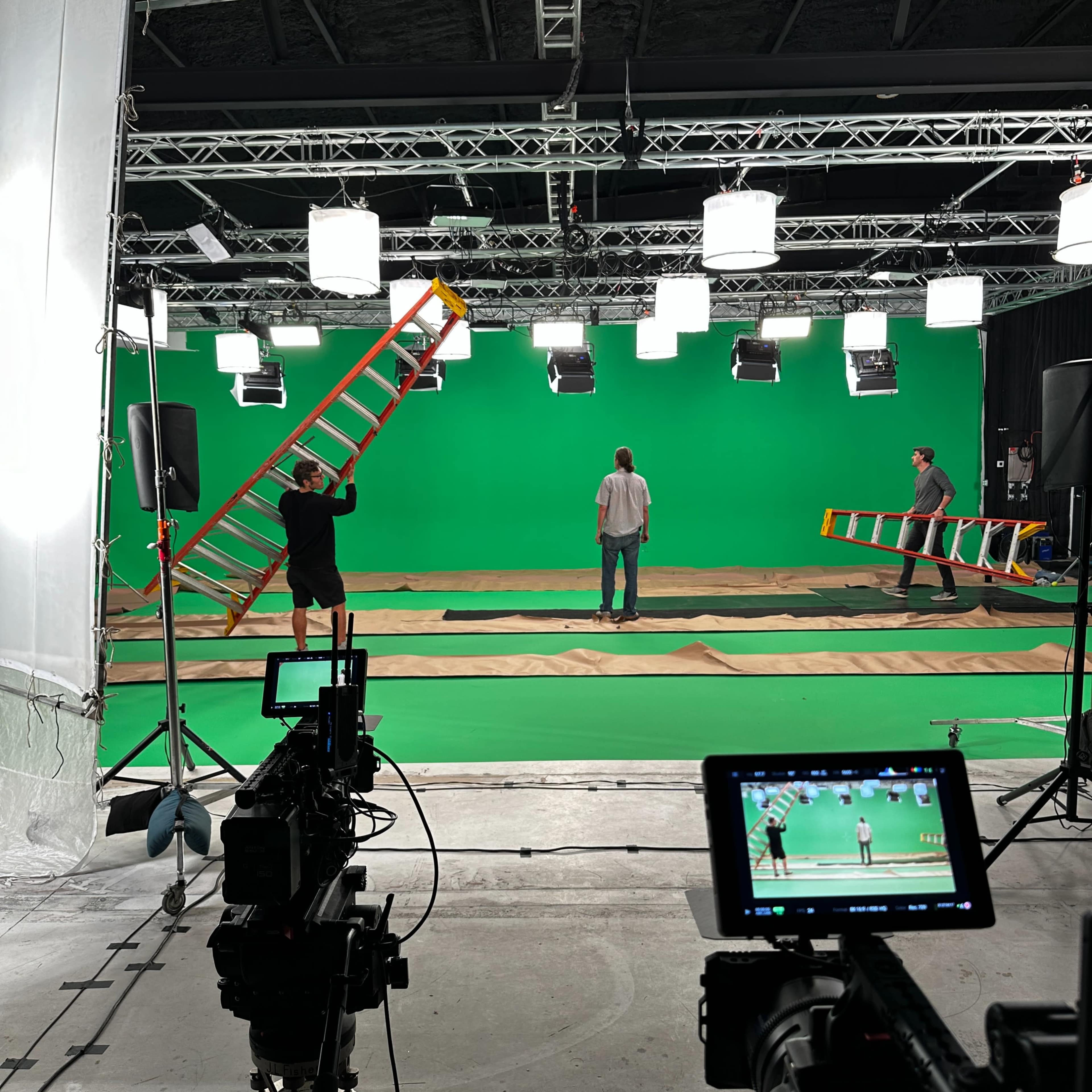 A video production studio is set up with a green screen backdrop, lighting equipment, and crew members preparing the scene.