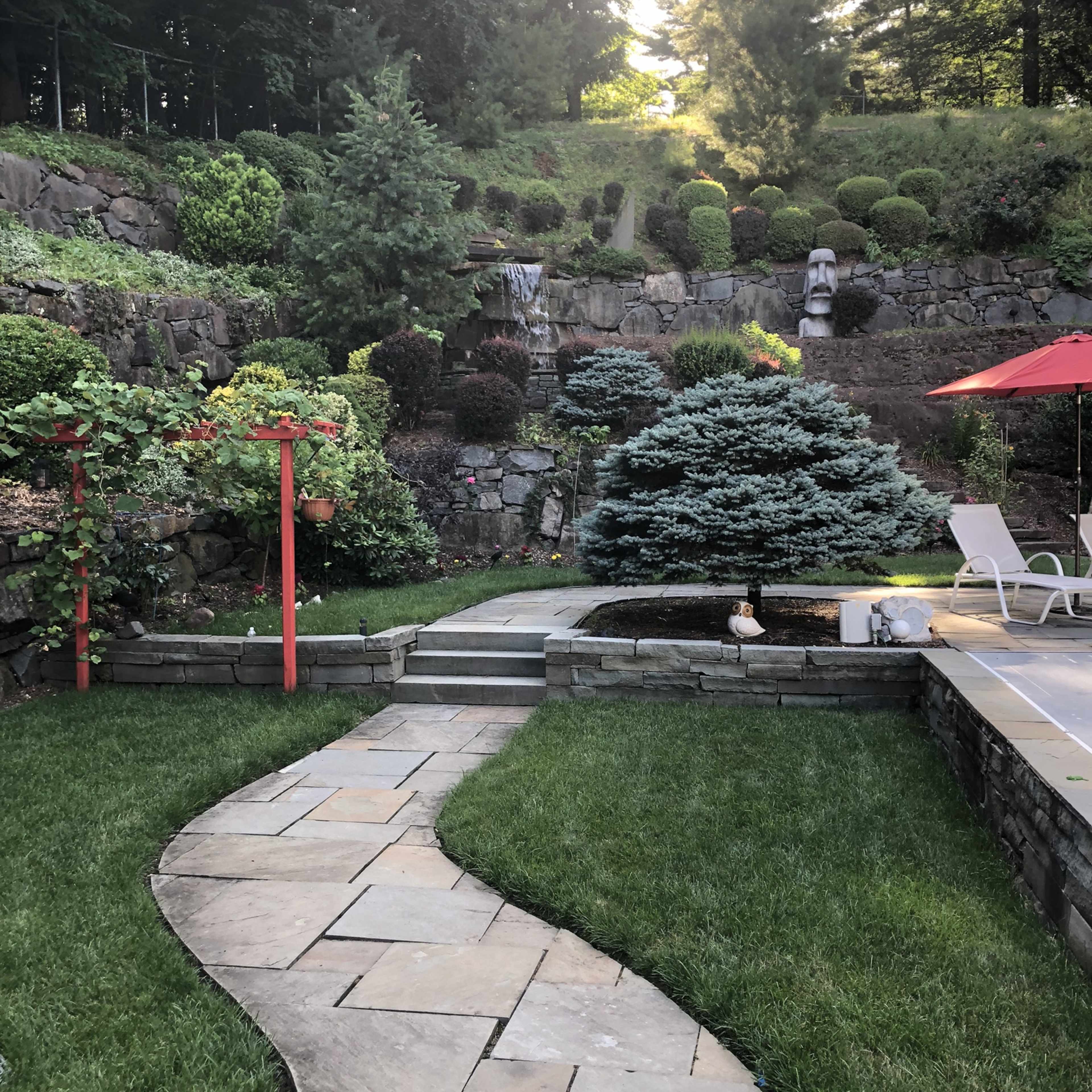 A landscaped garden with a stone pathway winding through greenery, leading to a small pond and a waterfall in a terraced setting.