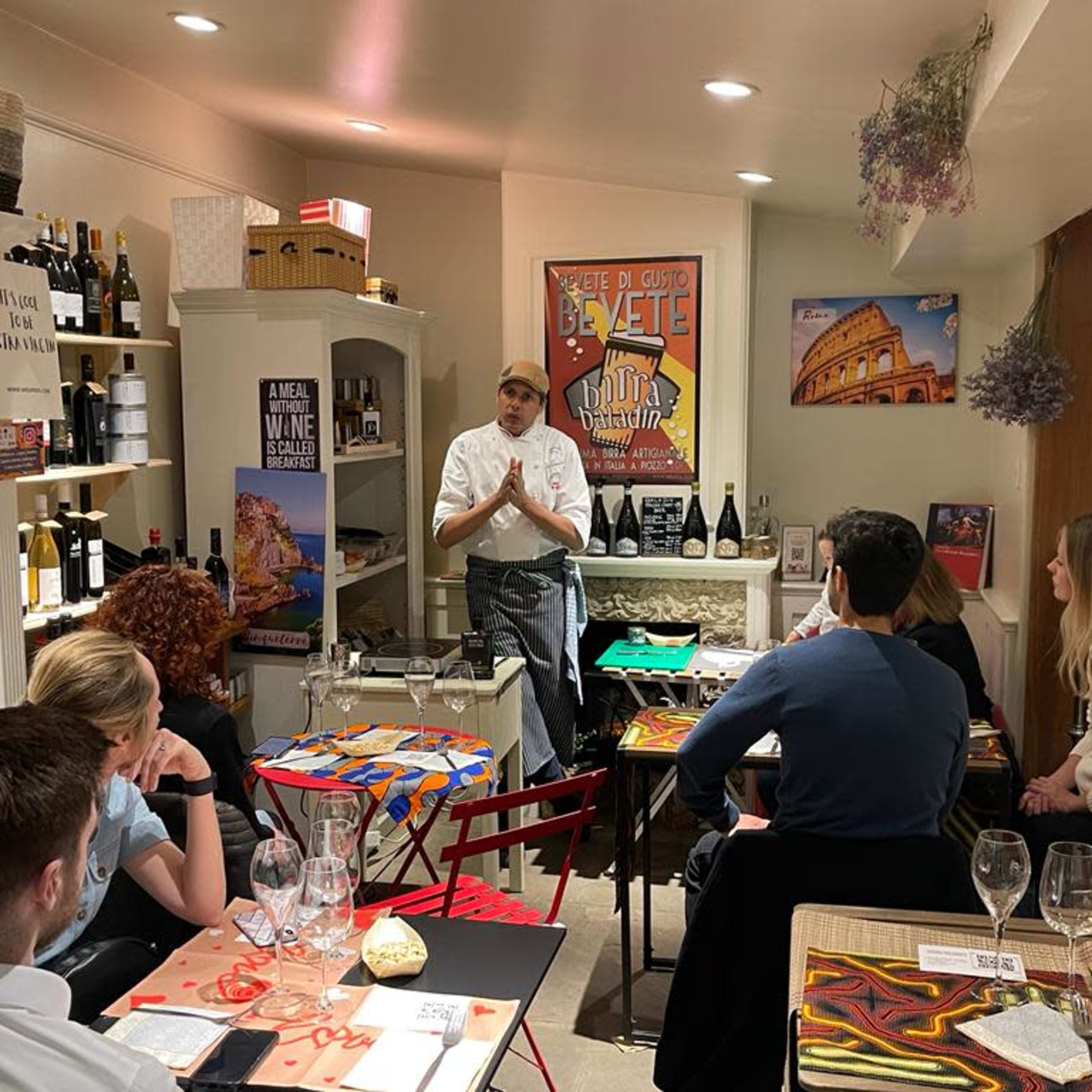 A chef stands in front of a small dining area, addressing a group of seated patrons amidst shelves of wine and food products.