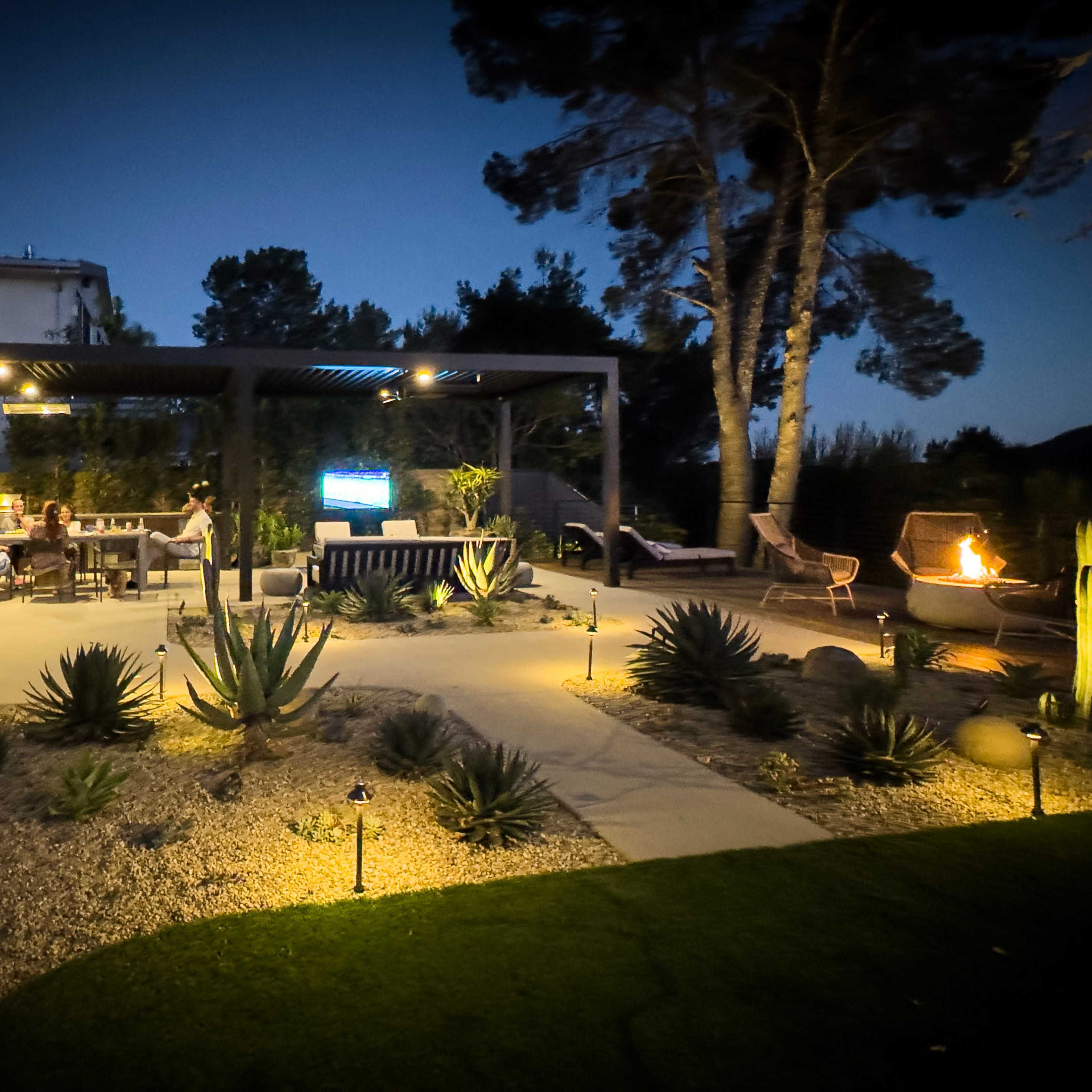 The scene shows a well-lit outdoor patio area with a dining table and fire pit surrounded by desert plants and trees at dusk.