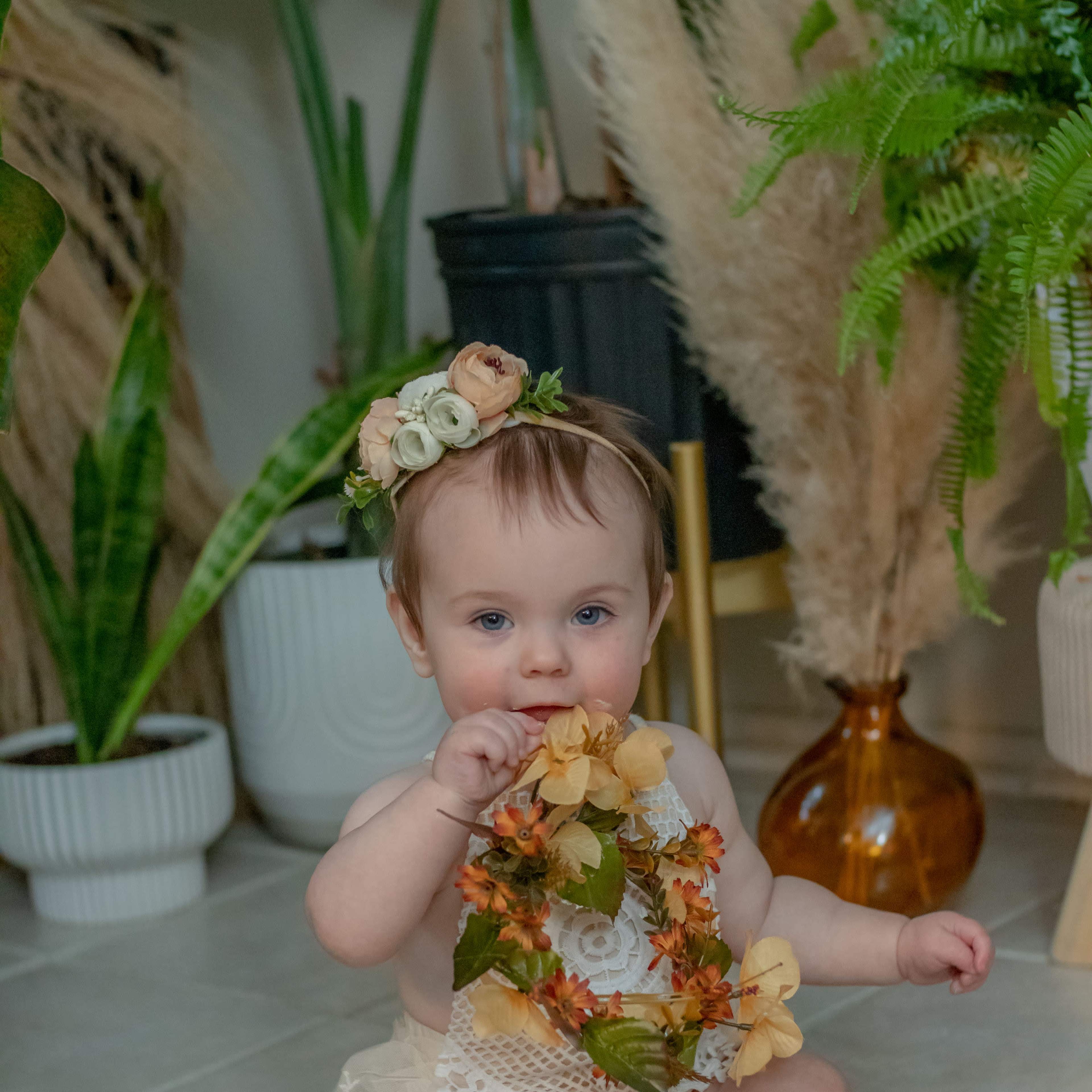 A baby sits on a tiled floor surrounded by plants, wearing a floral crown and holding a garland of flowers, with a cake in front of her.