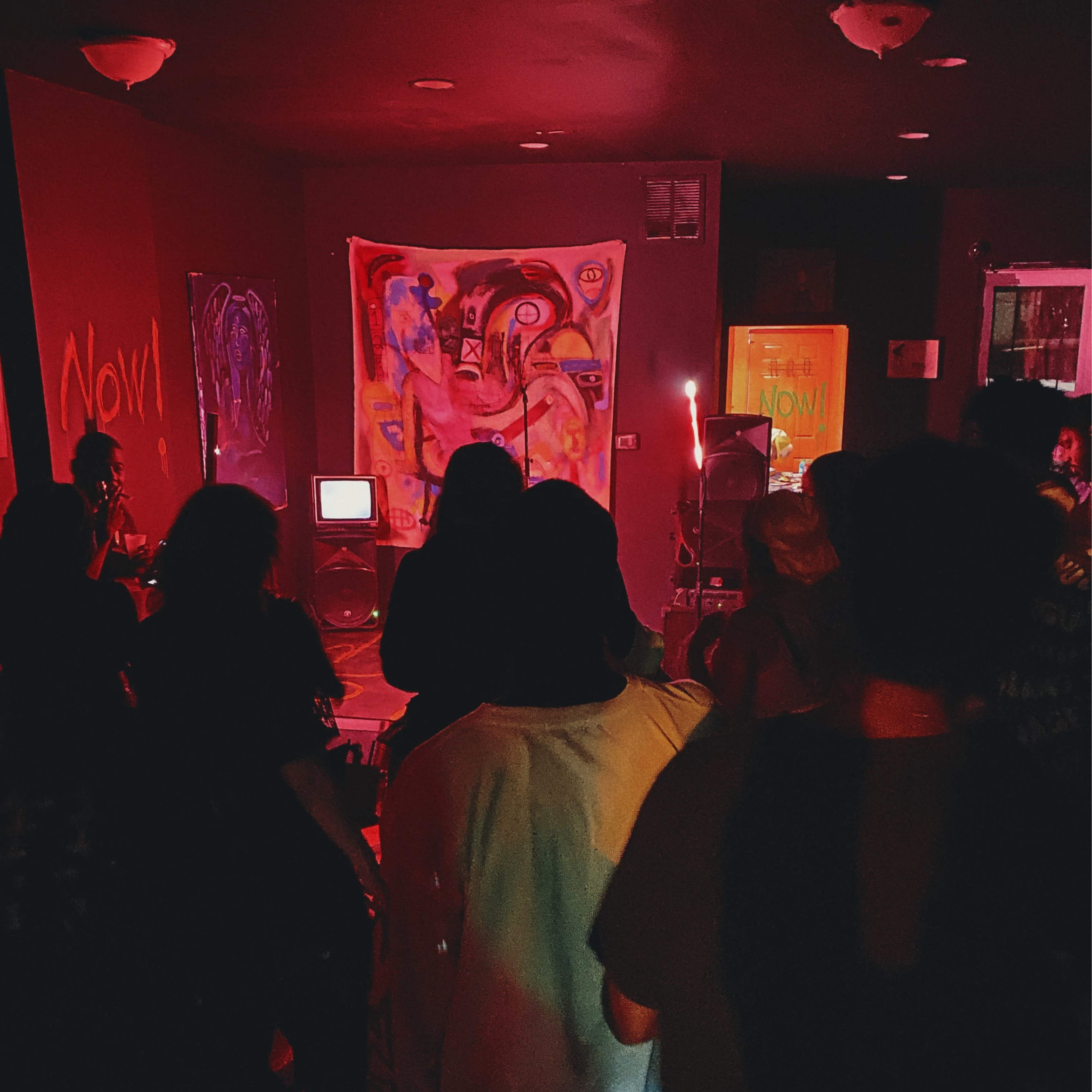 A group of people gathers in a dimly lit room with red lighting, facing a colorful wall art display while an old television set is positioned nearby.