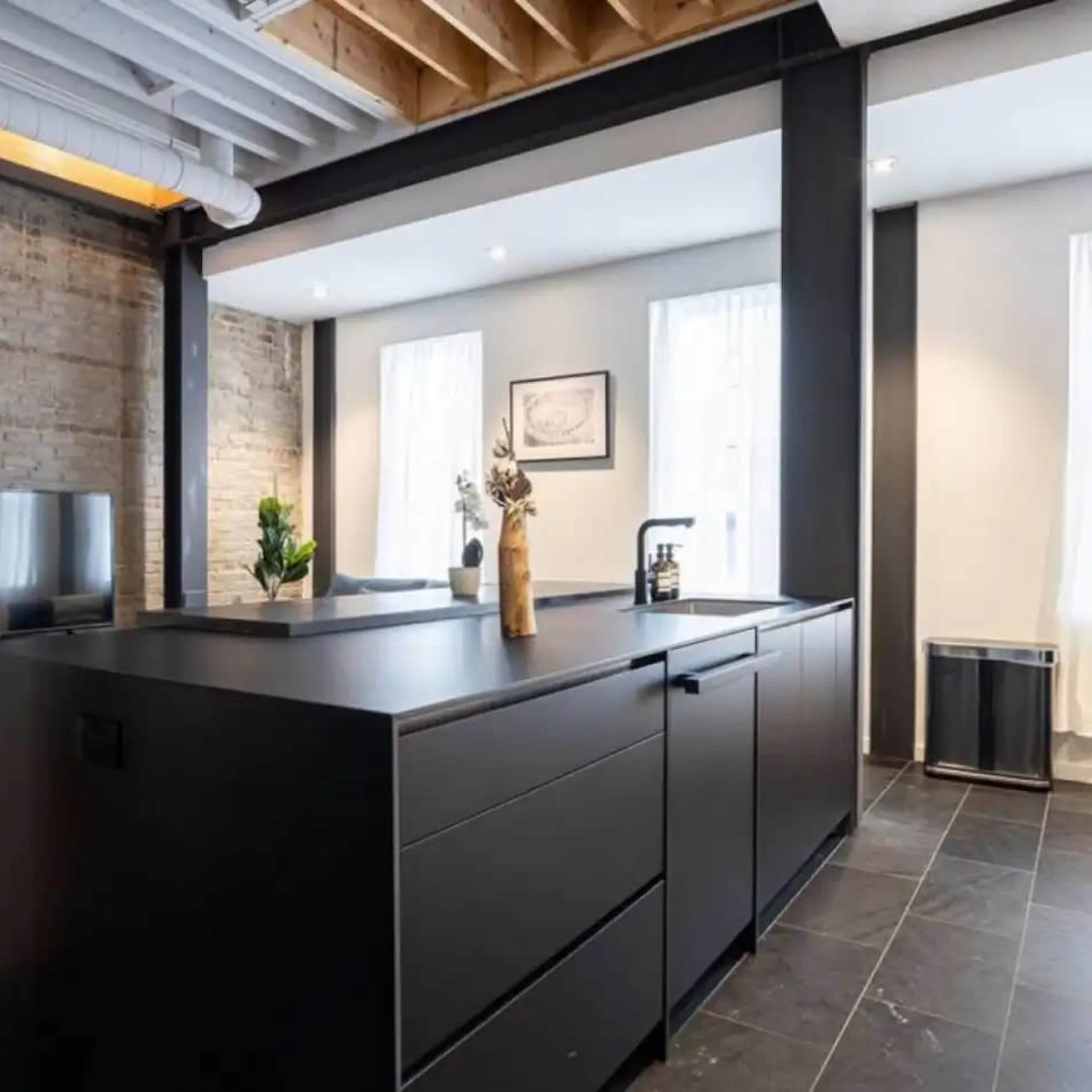 A modern kitchen with a large black island, stone walls, and large windows that let in natural light.
