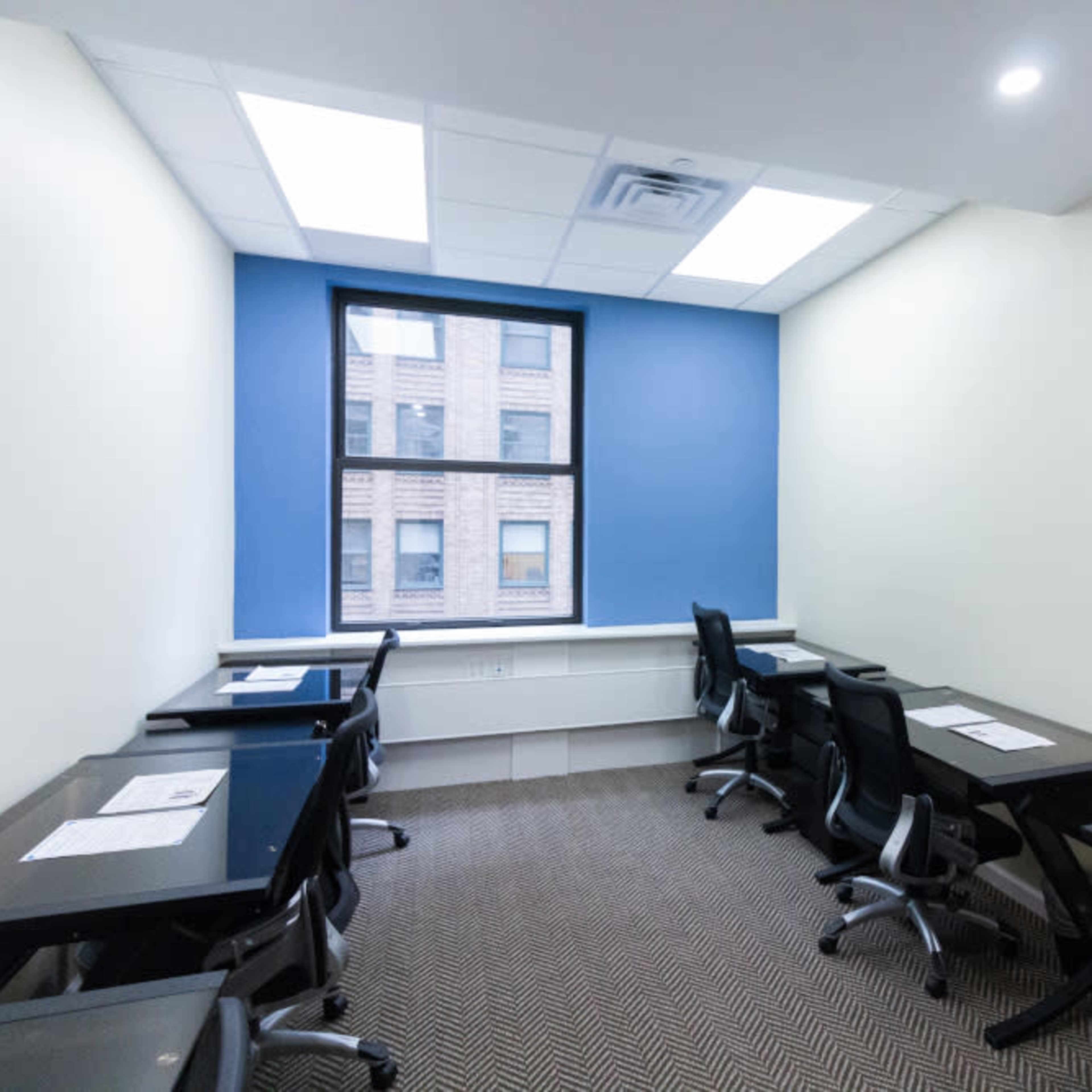 A small meeting room with several black chairs and desks arranged around a window with blue walls.