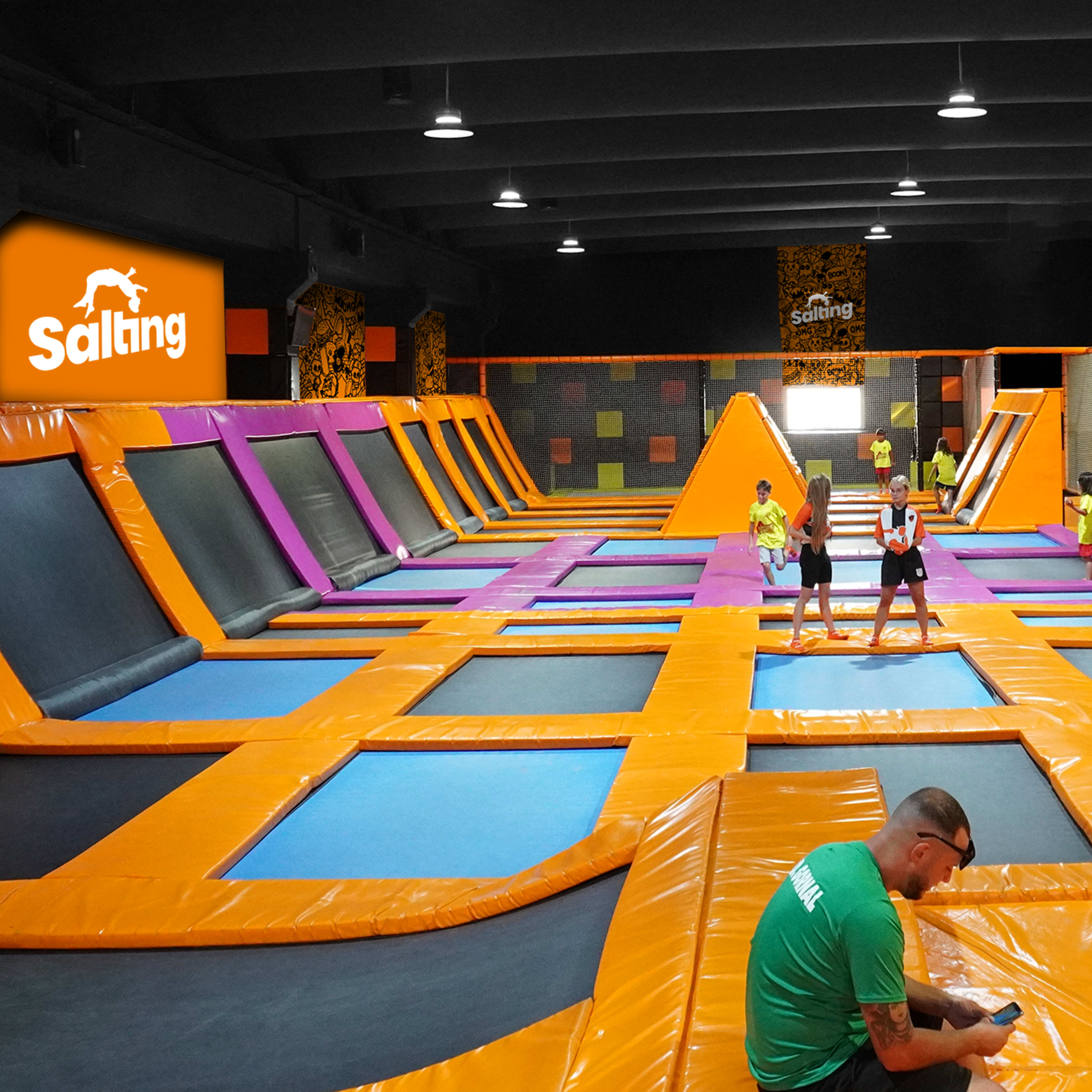 A vibrant indoor trampoline park with orange and black trampolines, colorful wall designs, and several children and adults jumping and exploring the area.