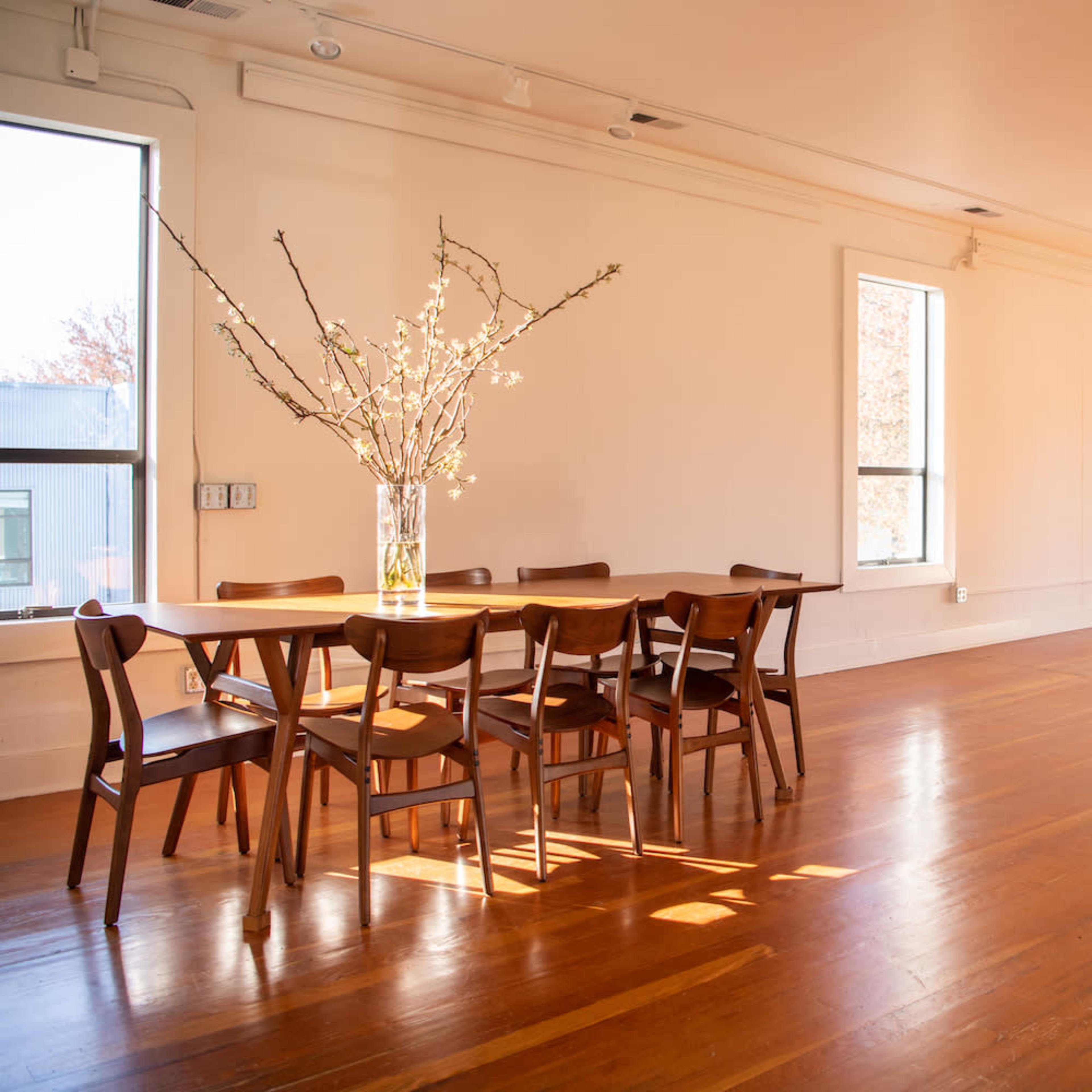 A spacious, brightly lit room features a wooden dining table surrounded by chairs, with large windows allowing sunlight to illuminate the hardwood floor.