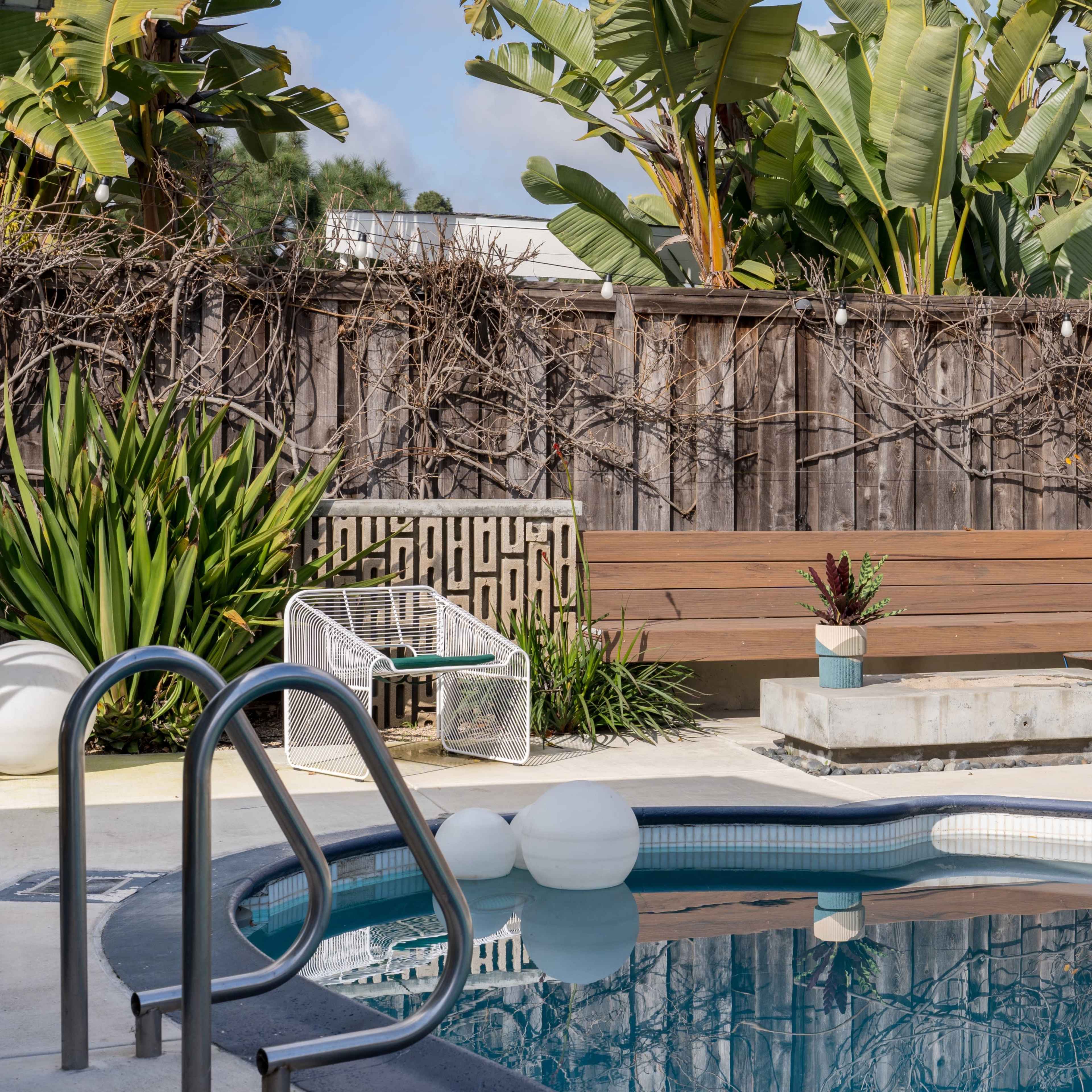 A swimming pool is surrounded by modern seating and lush green plants, with a wooden fence in the background.