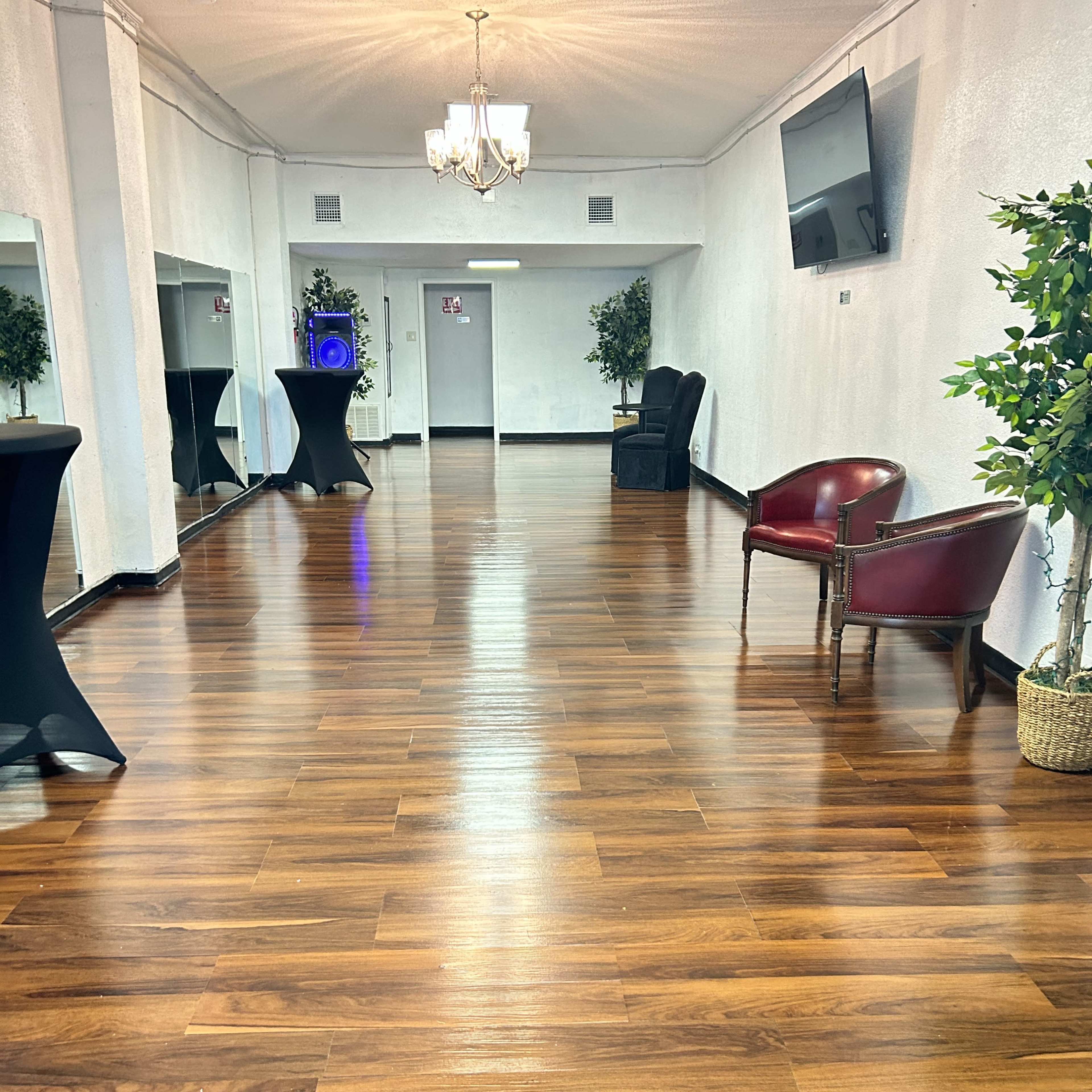 A spacious, well-lit room with polished wooden flooring, featuring a chandelier, mirrors along the walls, and a mix of seating arrangements including a chair and black cocktail tables.
