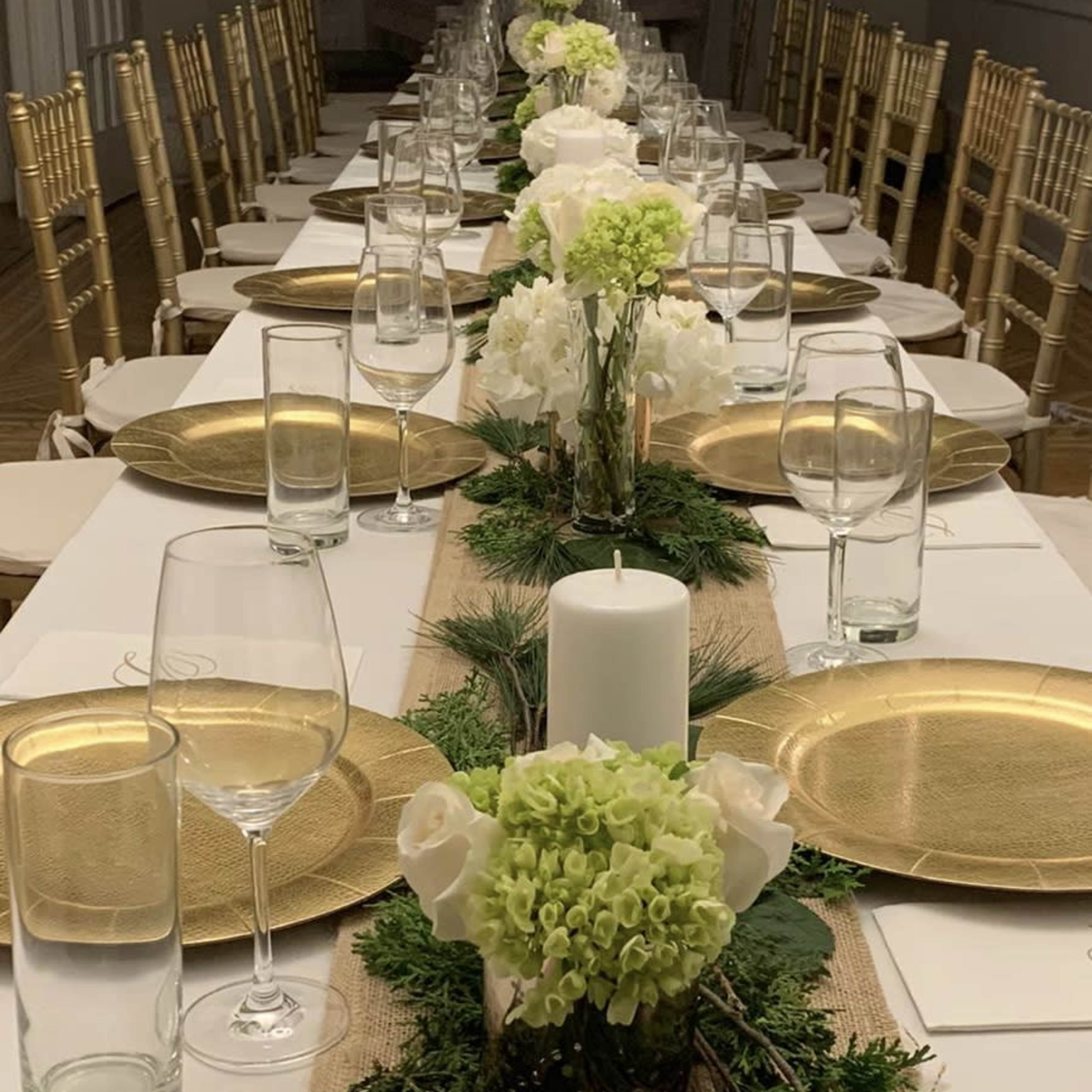 A long banquet table is set with gold-rimmed plates, glassware, and a centerpiece of white flowers and greenery, all arranged along a burlap runner.