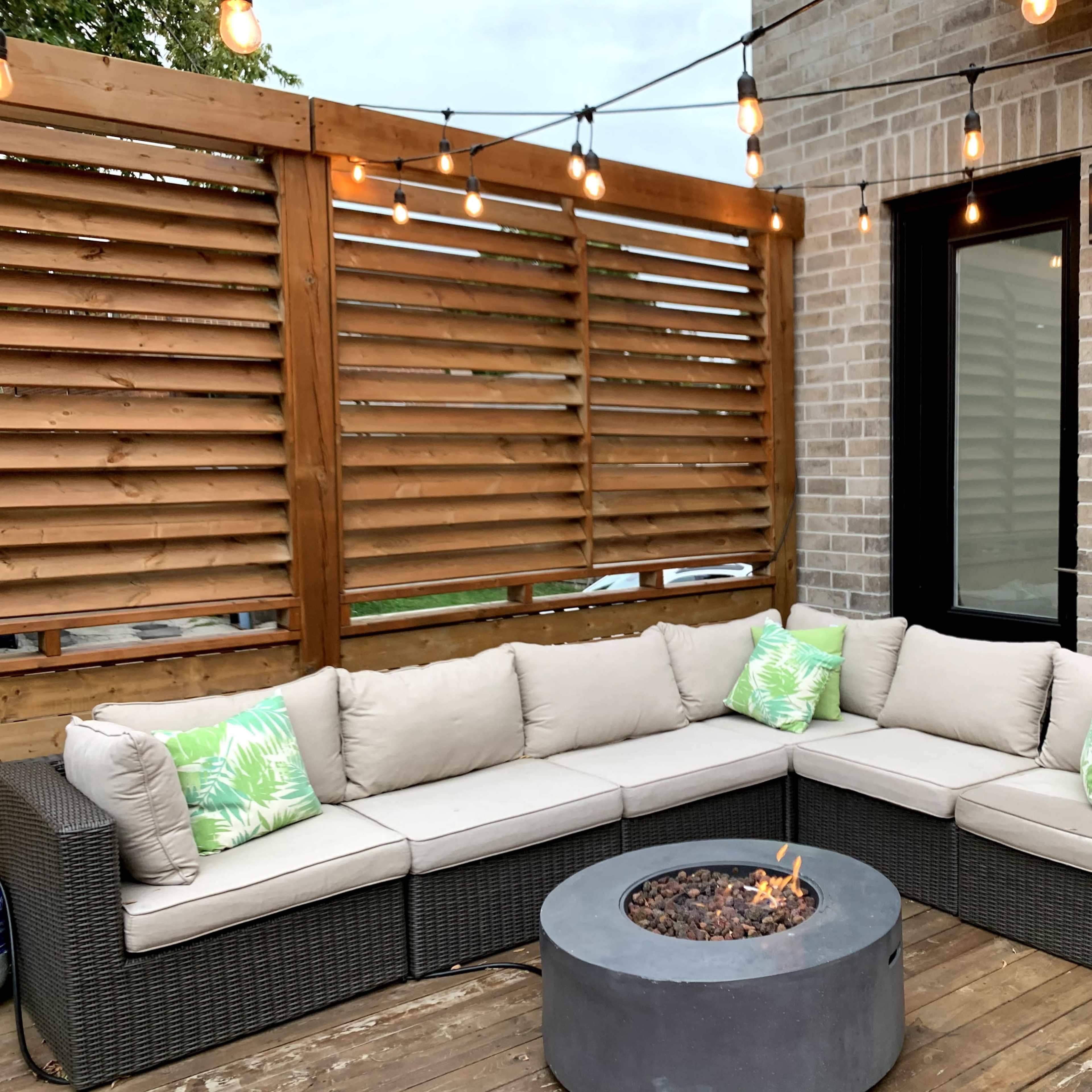 A corner seating area features a round fire pit surrounded by a sectional sofa on a wooden deck, with string lights overhead and a slatted wooden screen.