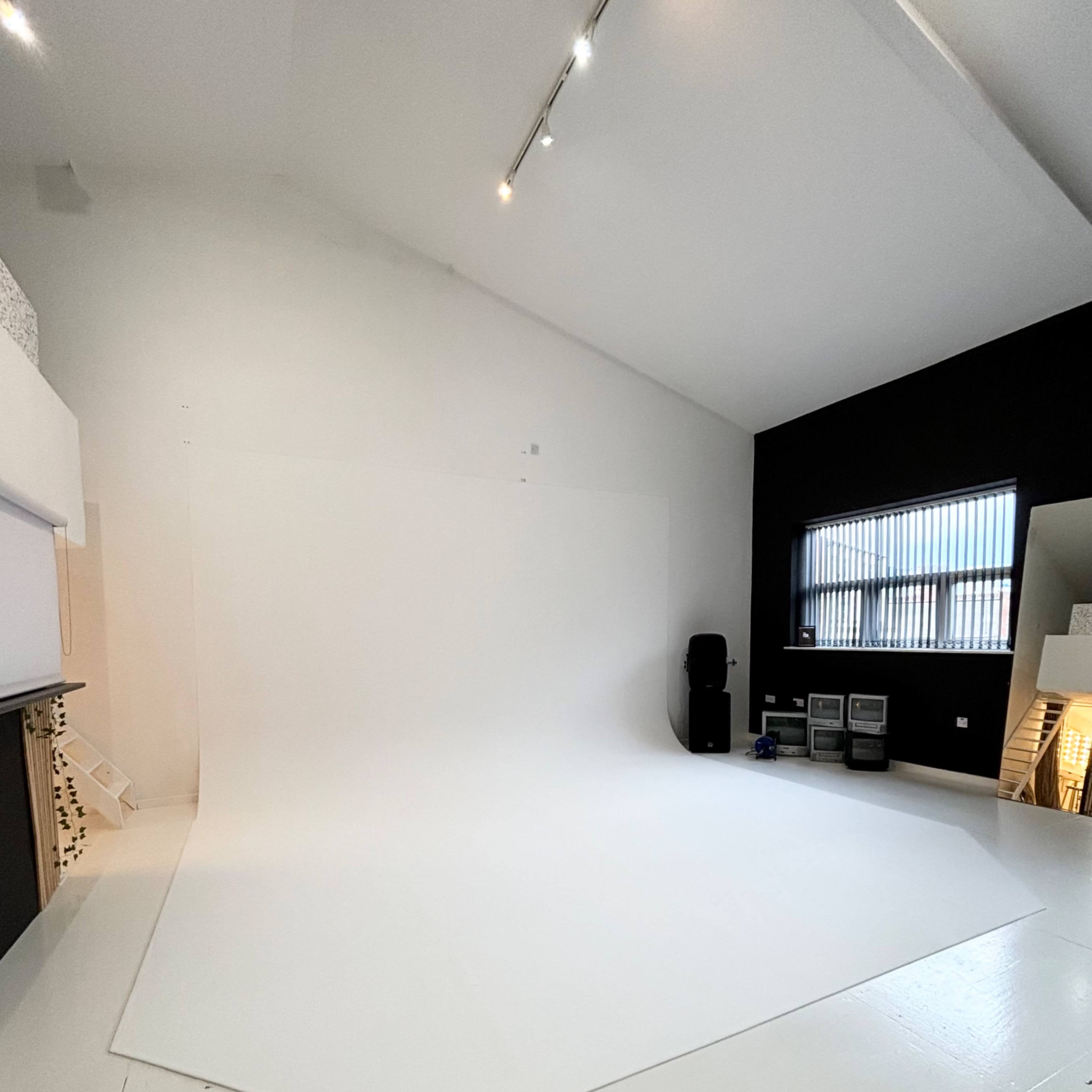 A spacious photography studio with a large white backdrop and a black wall, equipped with various studio equipment in the corner.
