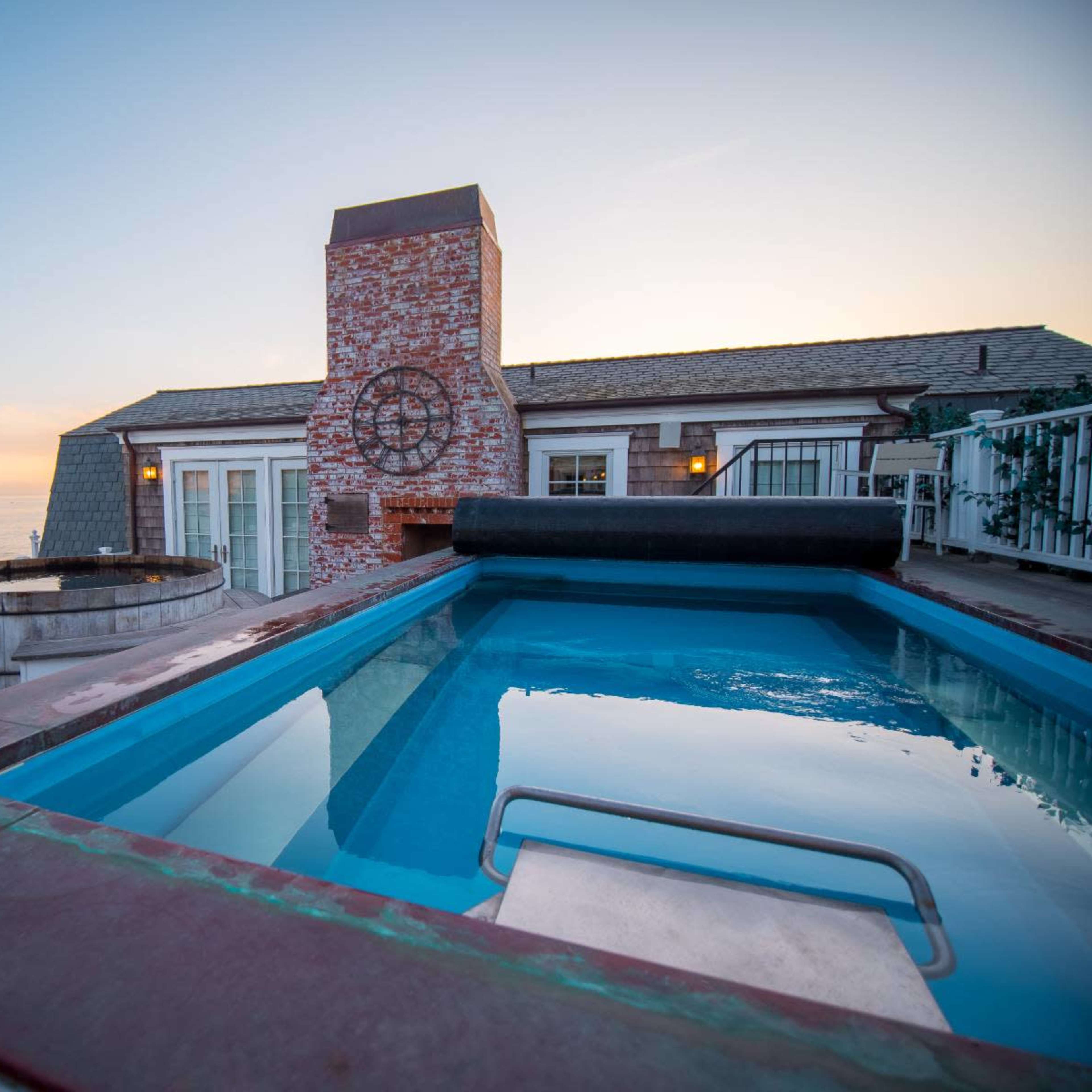A swimming pool with a built-in diving board is located next to a brick chimney and a hot tub, overlooking the ocean at sunset.
