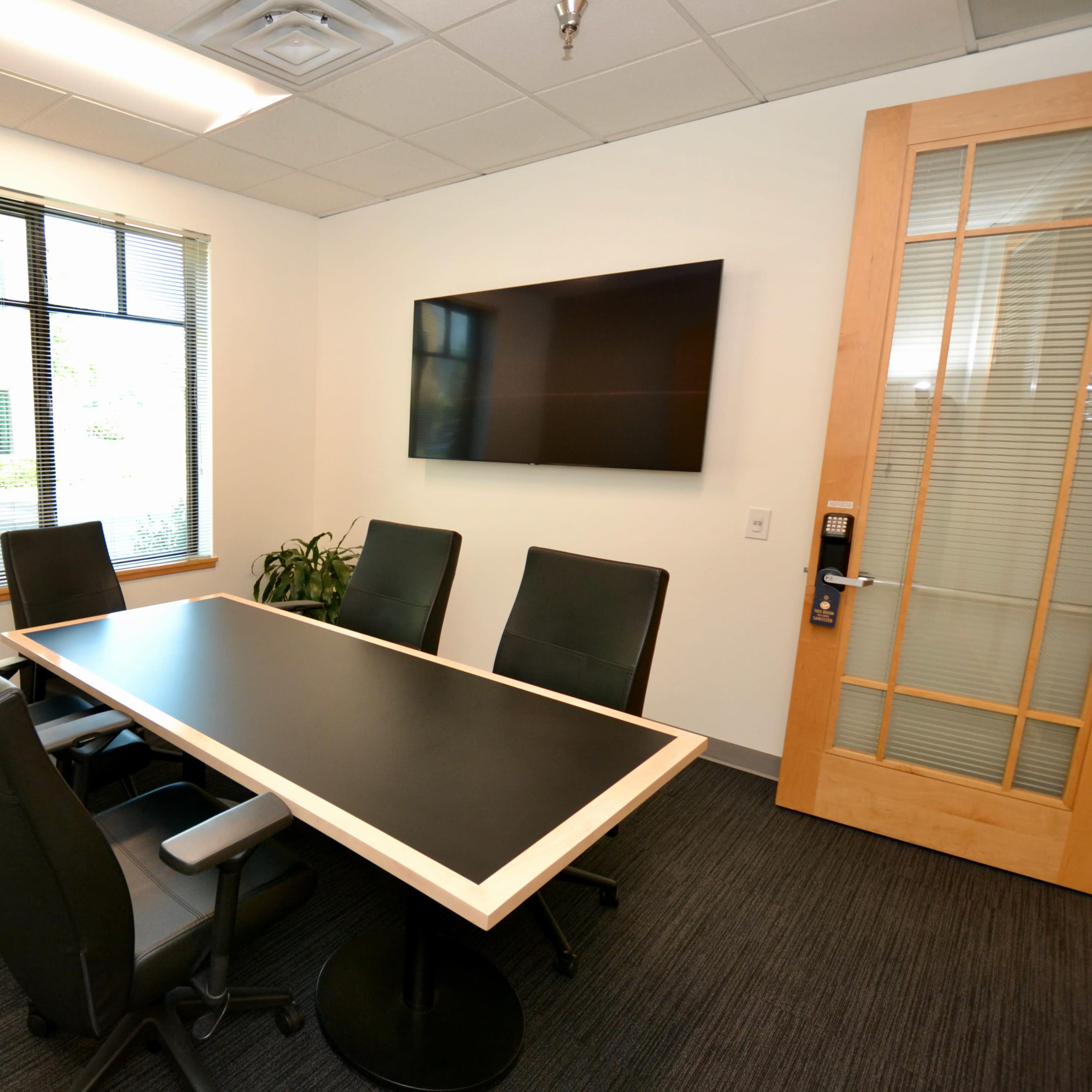 A small conference room with a black table, five chairs, a television on the wall, and a window with blinds.