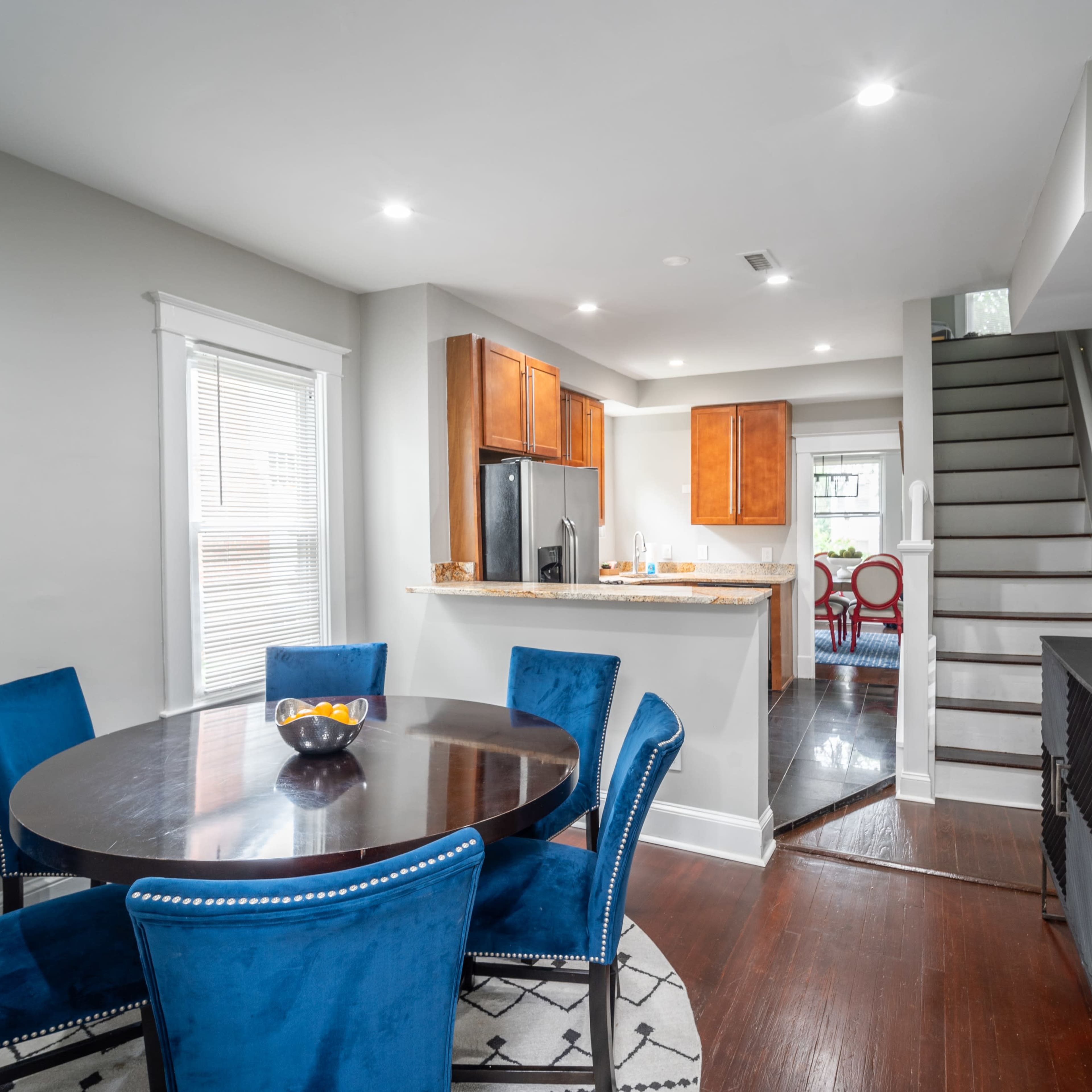 A modern dining area with a round table surrounded by blue upholstered chairs, adjacent to a kitchen and a staircase leading to an upper level.