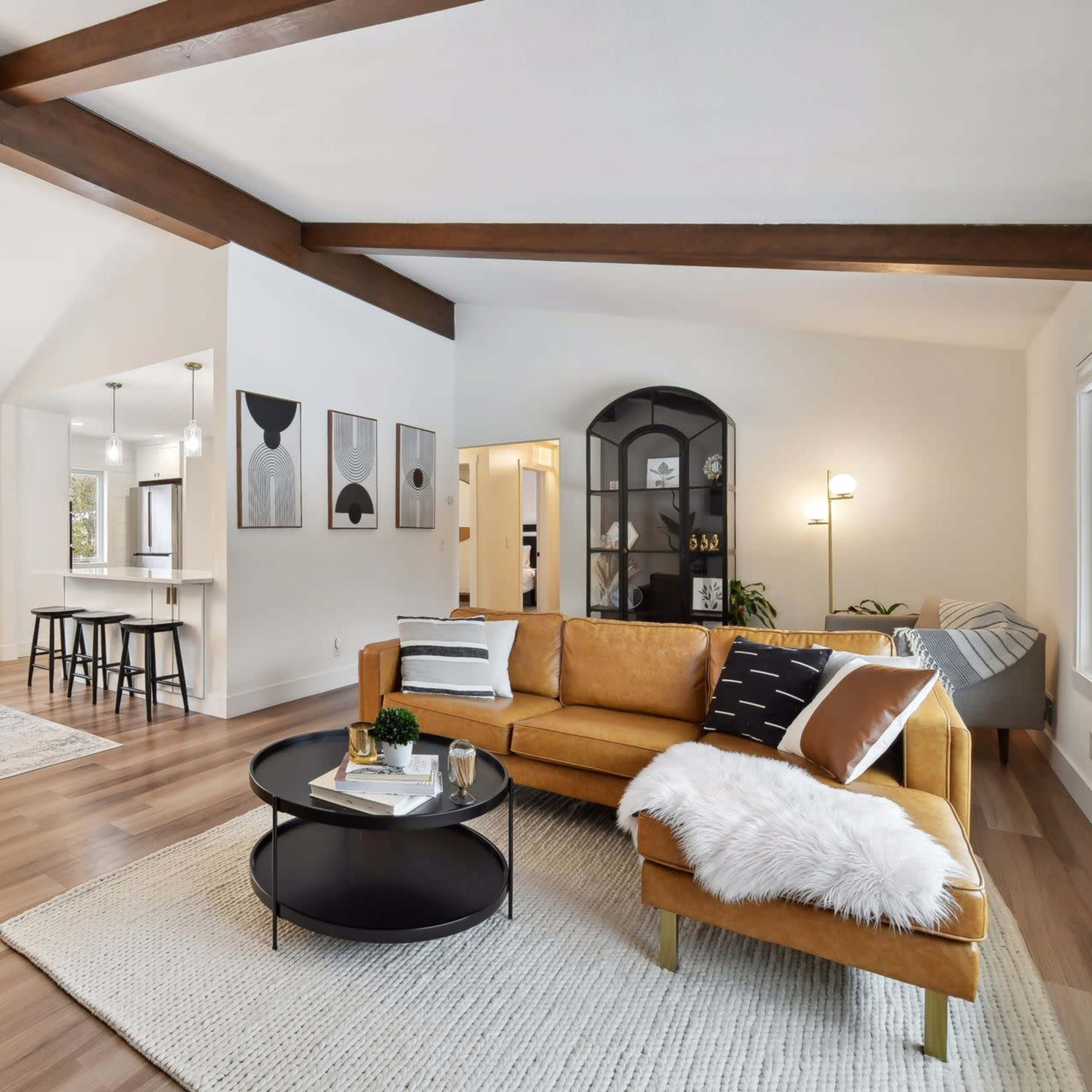 A bright, modern living room with a tan sofa, round coffee table, and wooden beams, connected to a dining area with a black table and chairs.