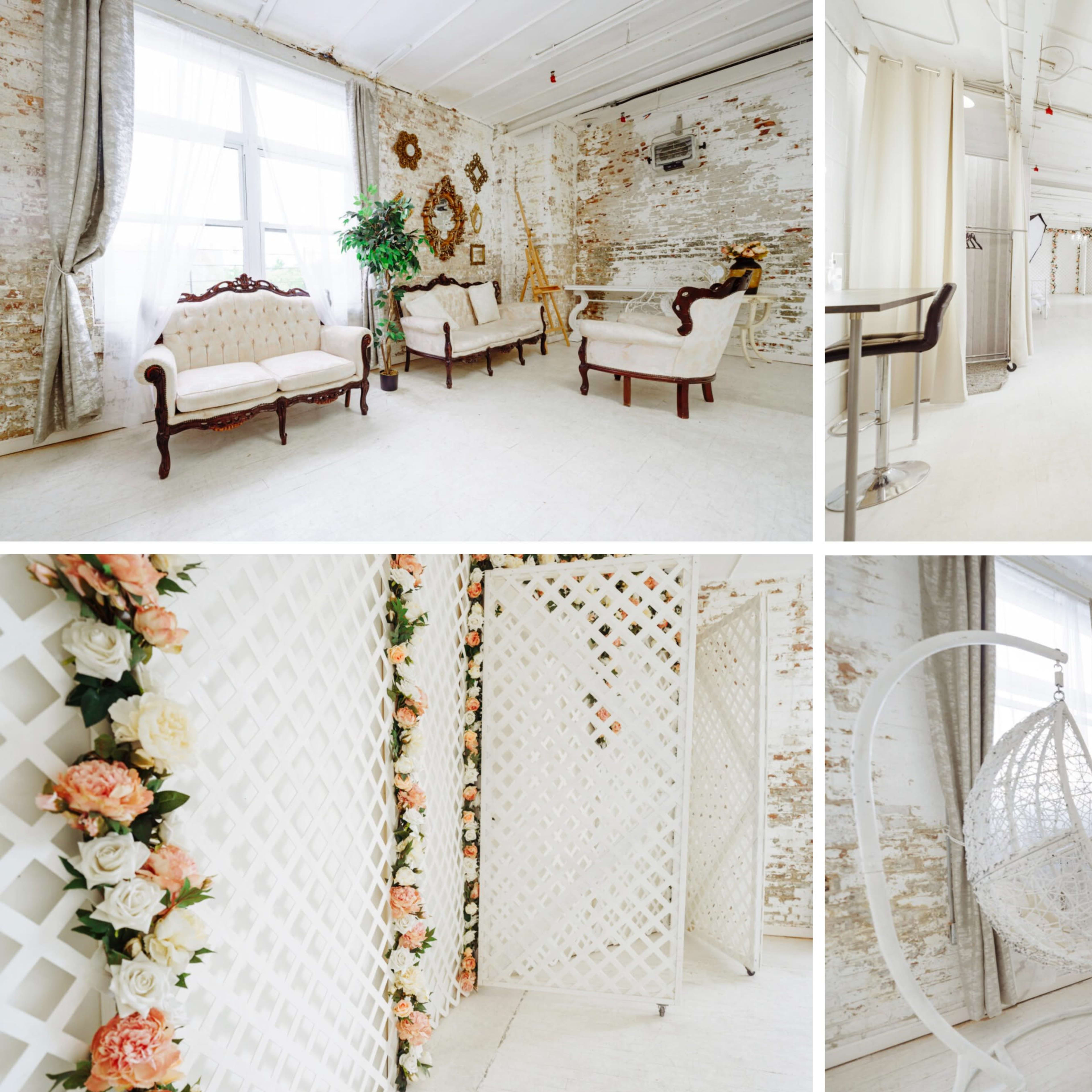 A bright, airy interior space featuring white walls, vintage furniture, decorative mirrors, and floral arrangements.