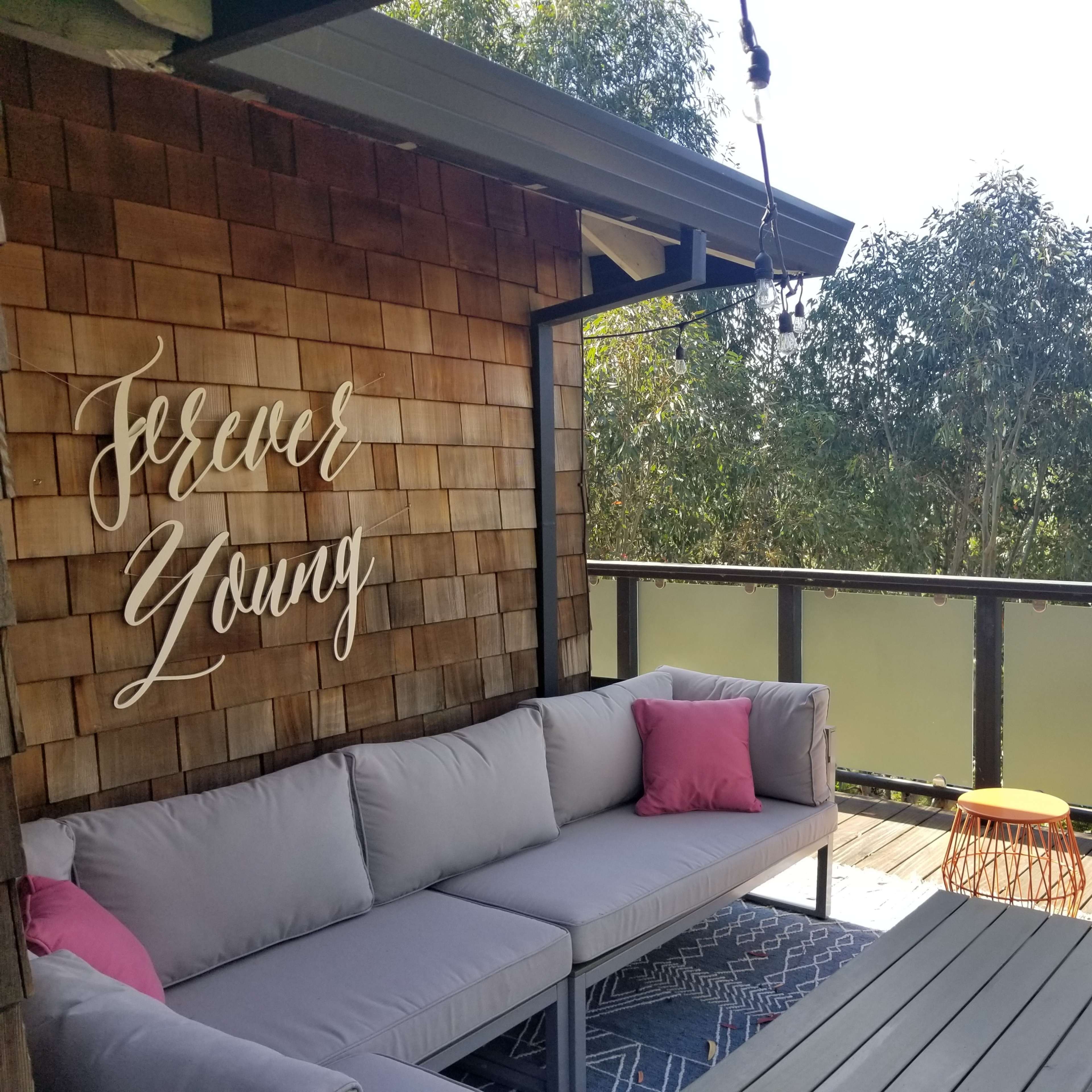 A cozy outdoor seating area features a gray sofa with pink cushions and two orange tables, against a backdrop of greenery and a wooden wall displaying the words "Forever Young."