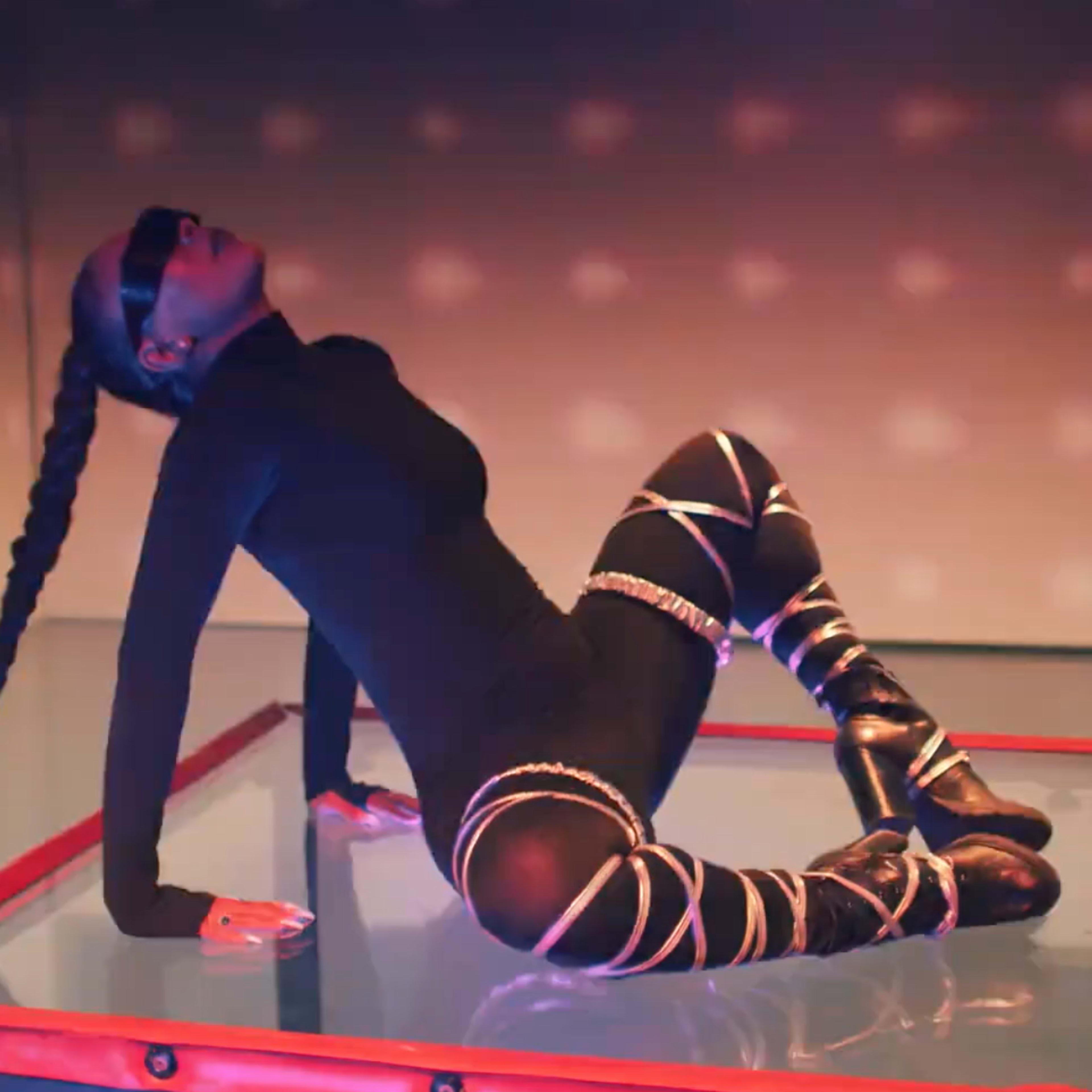 A person in a black outfit with long braids poses on a raised platform surrounded by soft lighting.
