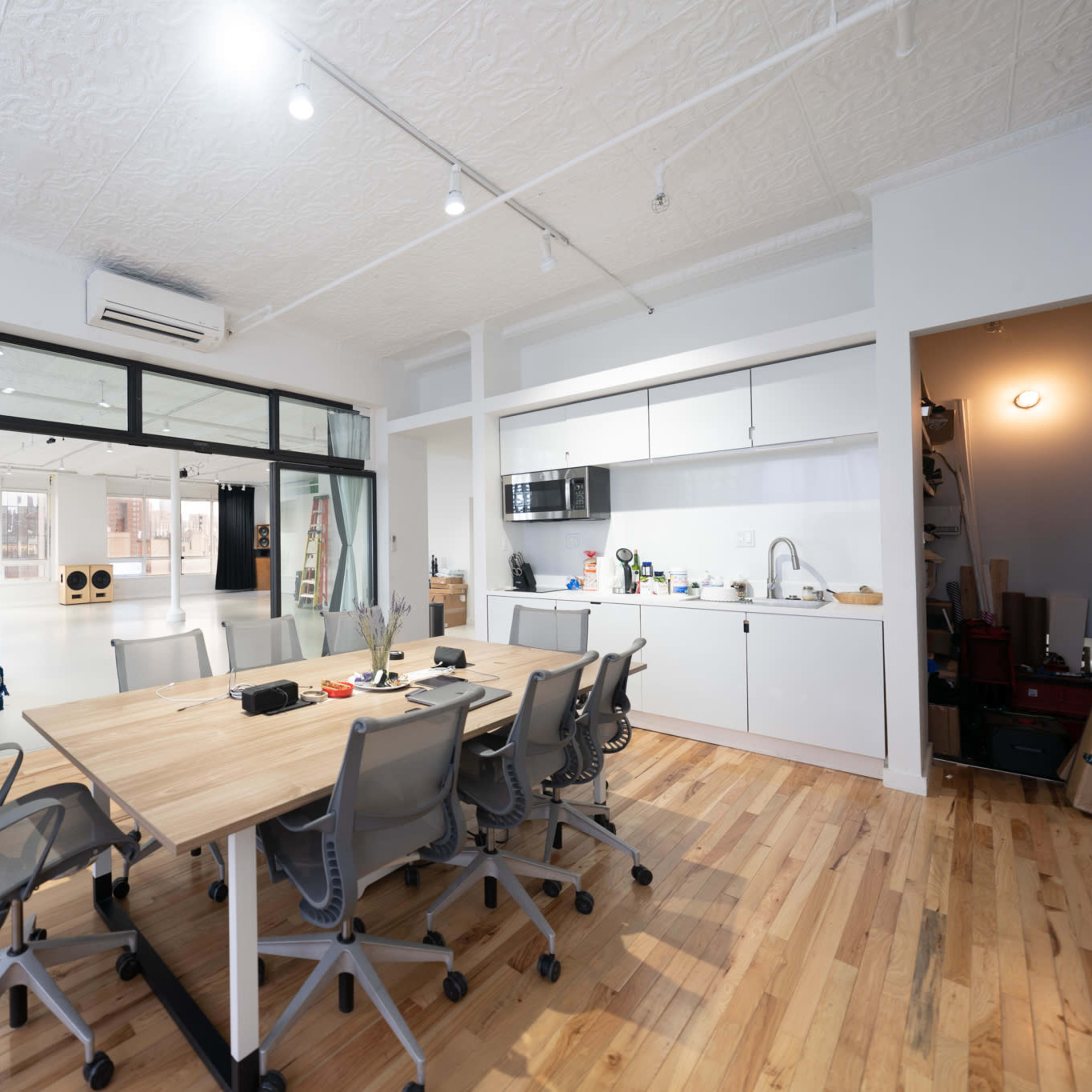 A spacious office features a large wooden table surrounded by ergonomic chairs, with a kitchenette and glass doors leading to a separate area.