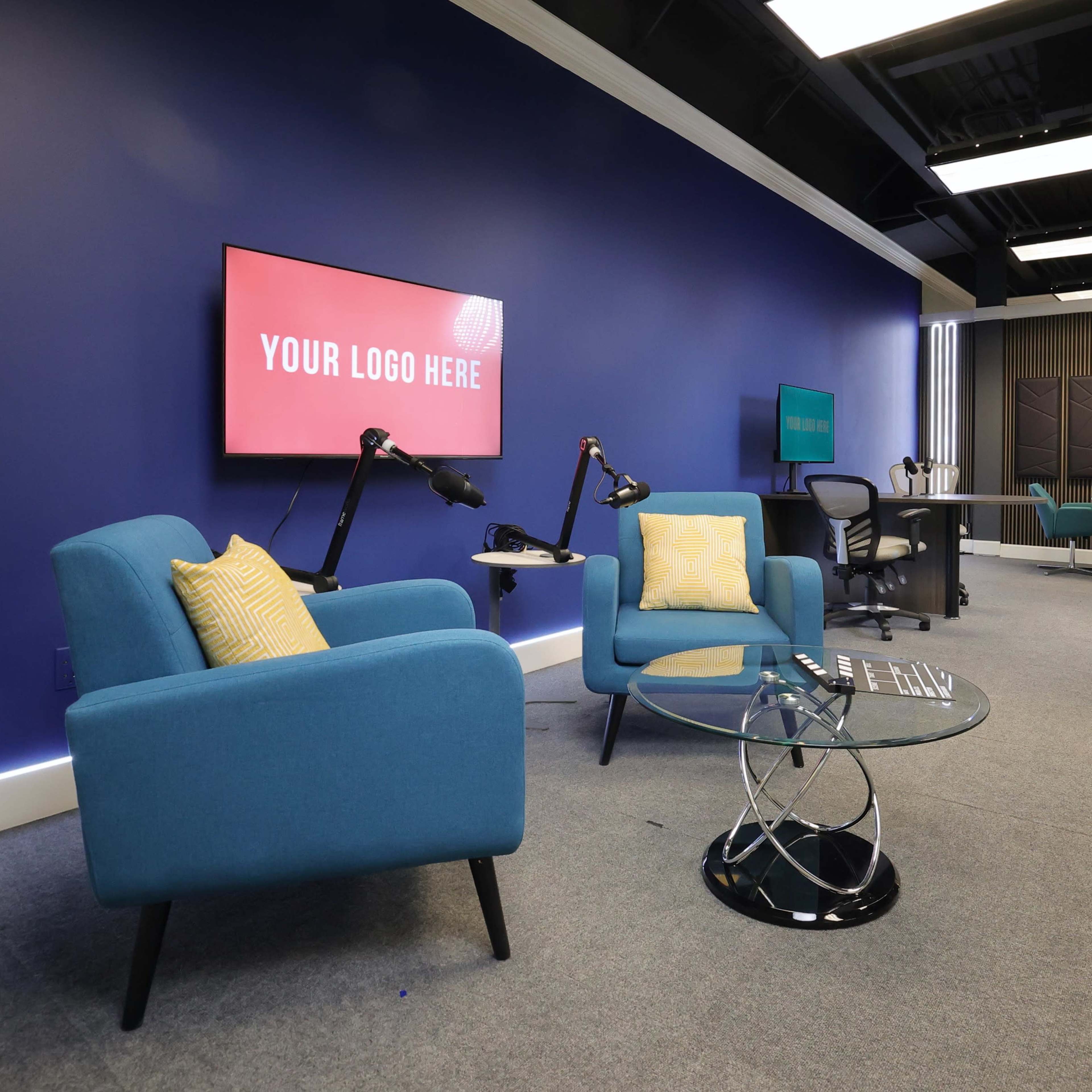 A modern studio space featuring two light blue chairs, a glass coffee table, a large screen with a placeholder for a logo, and various equipment for filming.