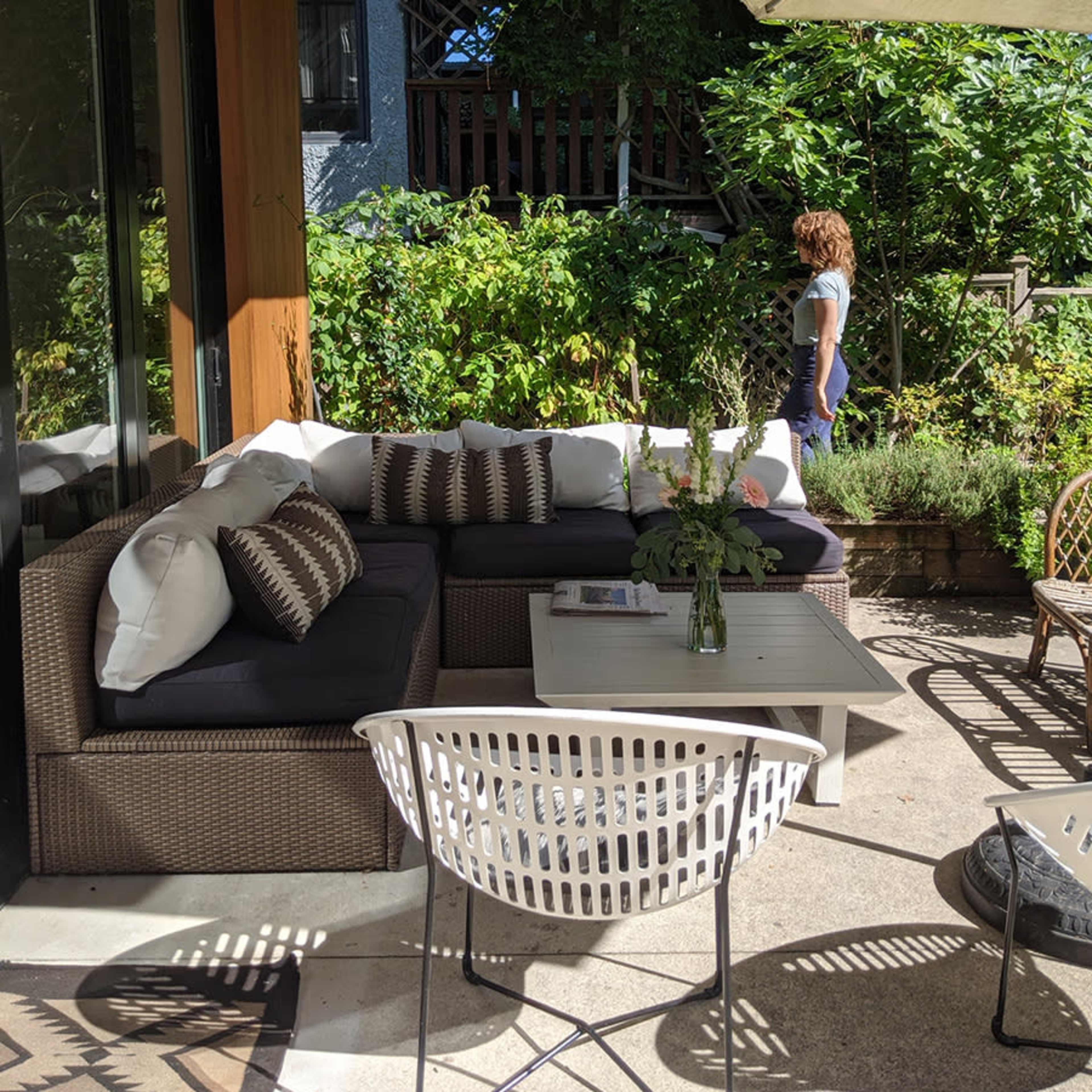 An outdoor seating area features a sectional sofa with pillows, a coffee table, and a woman walking in the background amidst greenery.