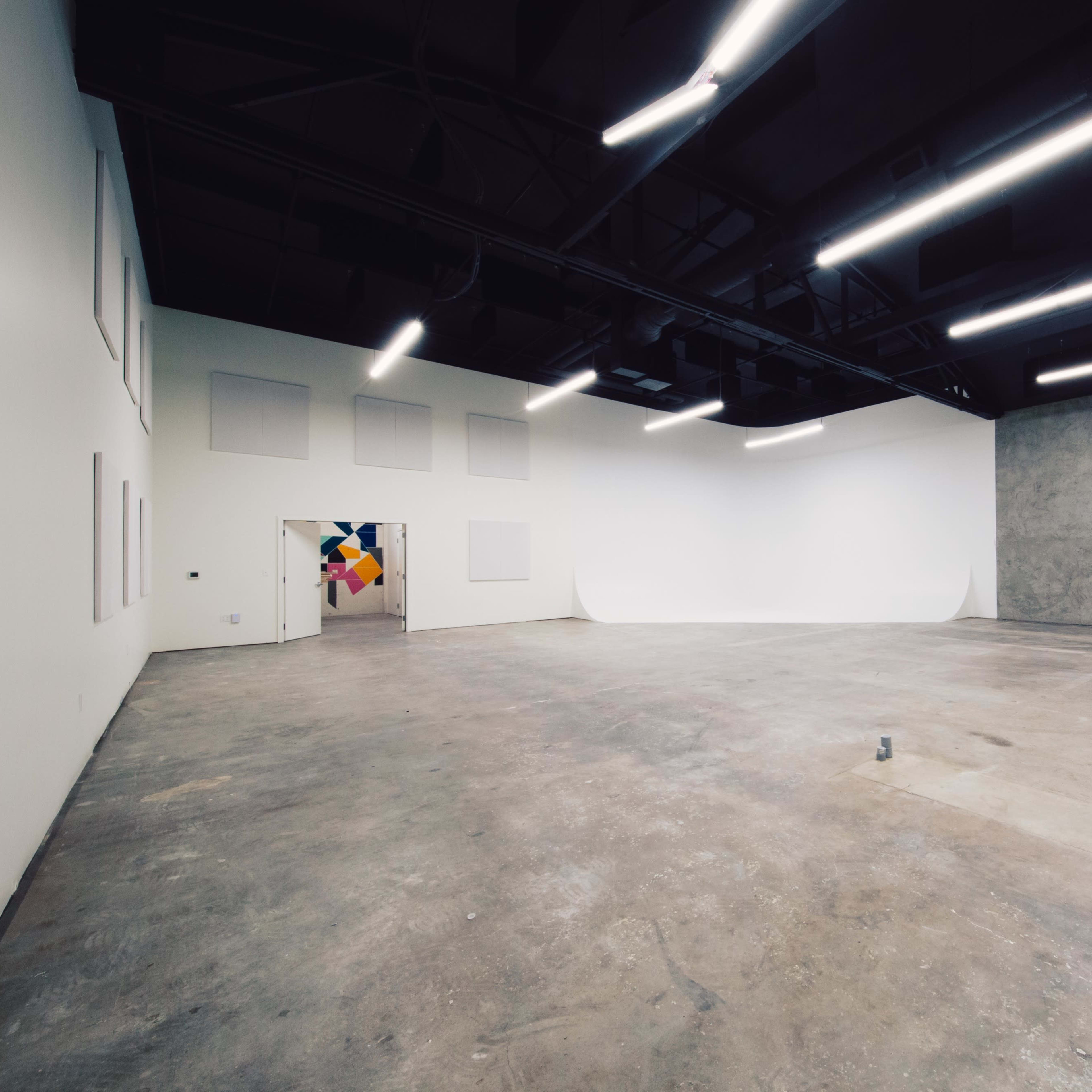 A spacious, empty gallery with white walls, a concrete wall on one side, and overhead fluorescent lighting.