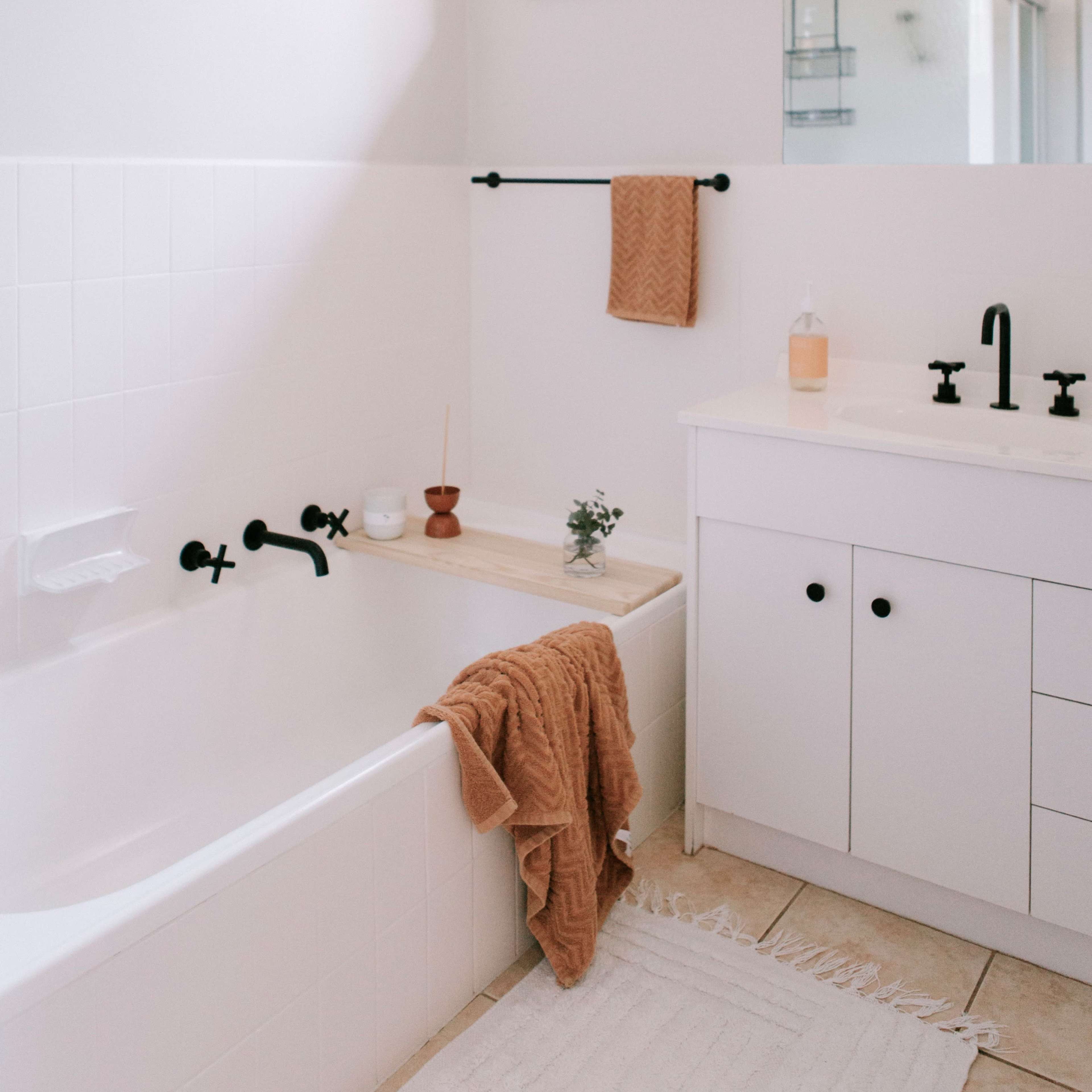 A bright, minimalist bathroom features a white bathtub with black fixtures, a neutral-toned towel draped over the side, and a small plant on the counter.