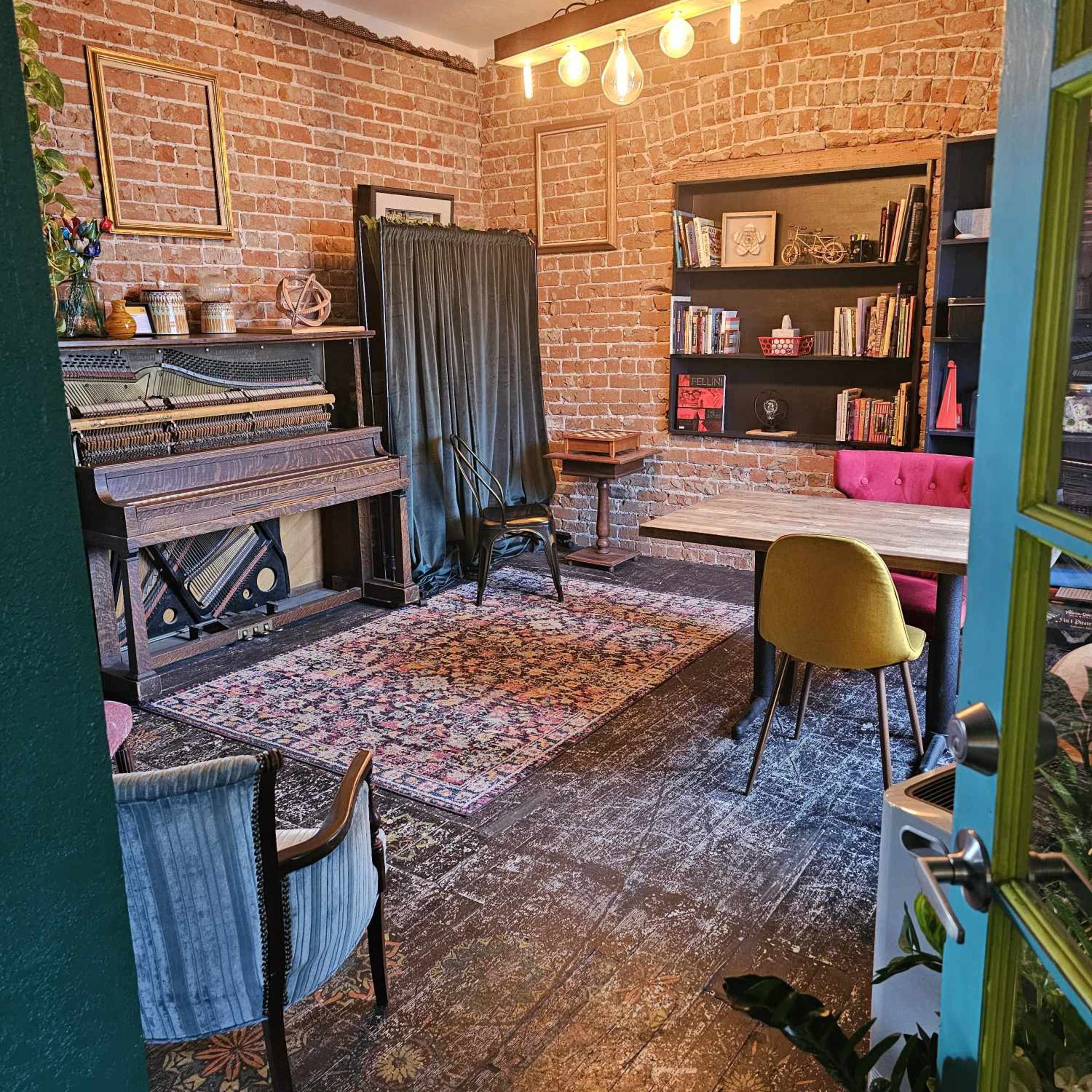 An interior room featuring a vintage piano, a reading area with bookshelves, and a mix of antique and modern furniture against a brick wall.