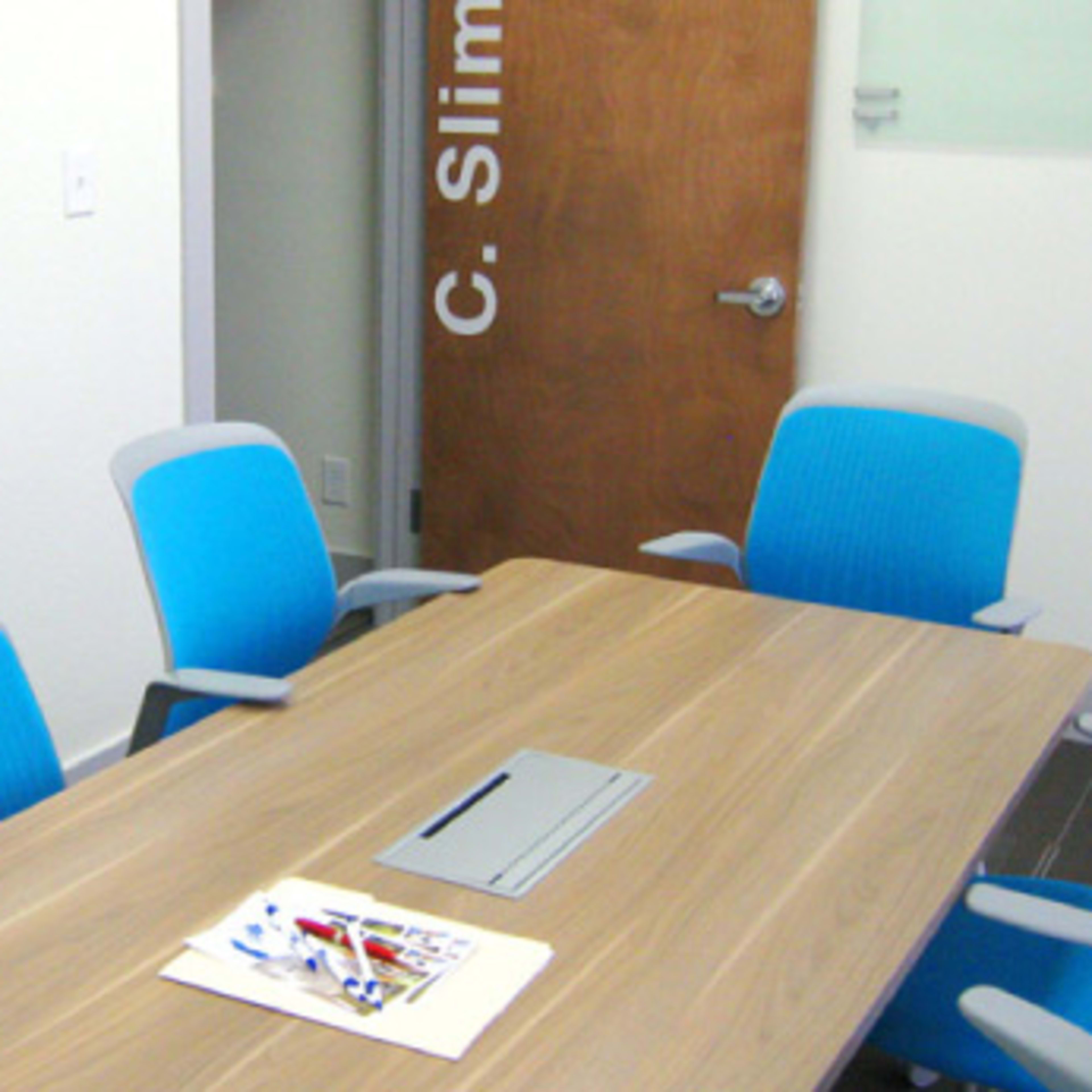 A conference room features a rectangular wooden table surrounded by blue and gray chairs, with a door labeled "C. Slim" visible in the background.