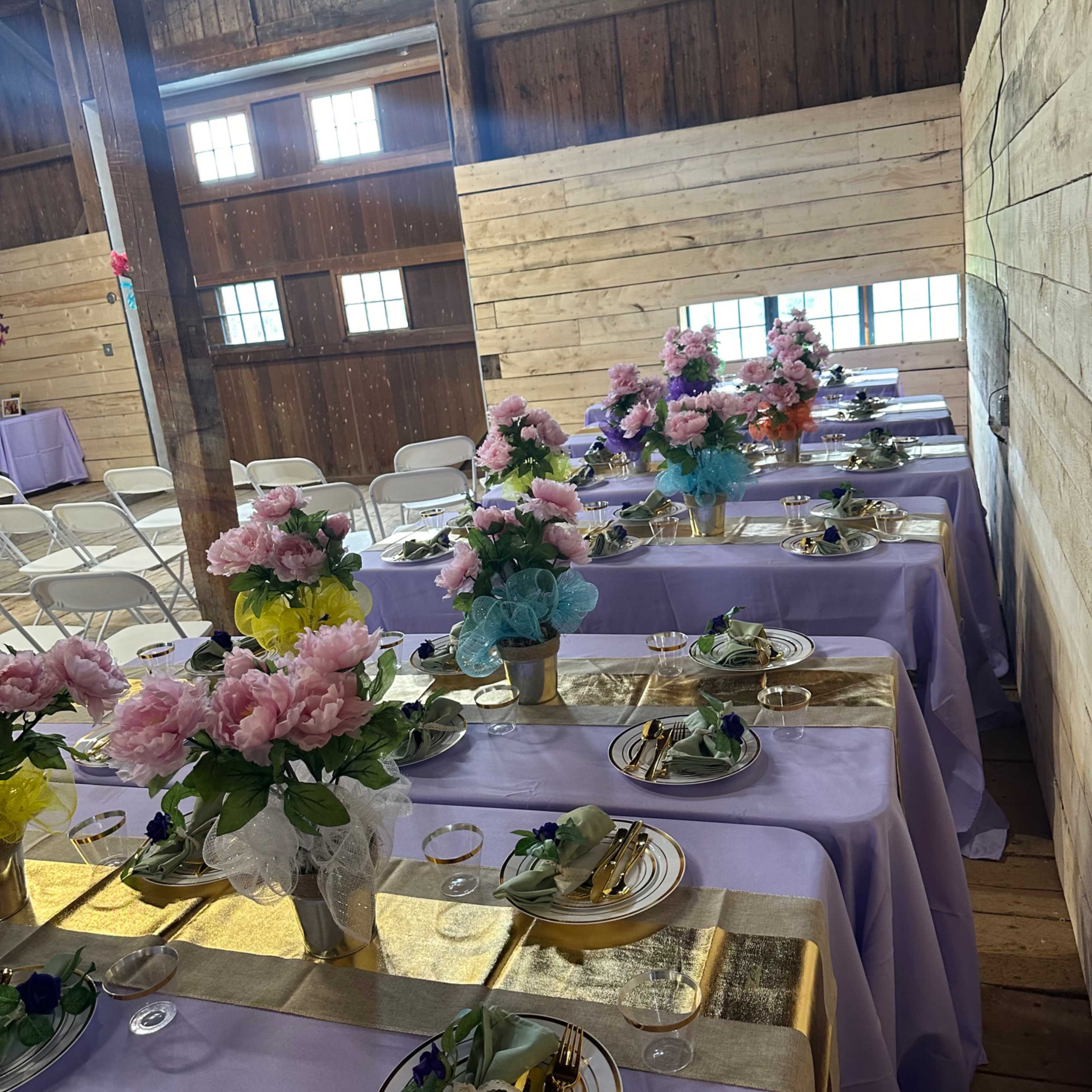 A decorated barn interior with tables set with floral centerpieces, tablecloths, and dinnerware arranged for an event.