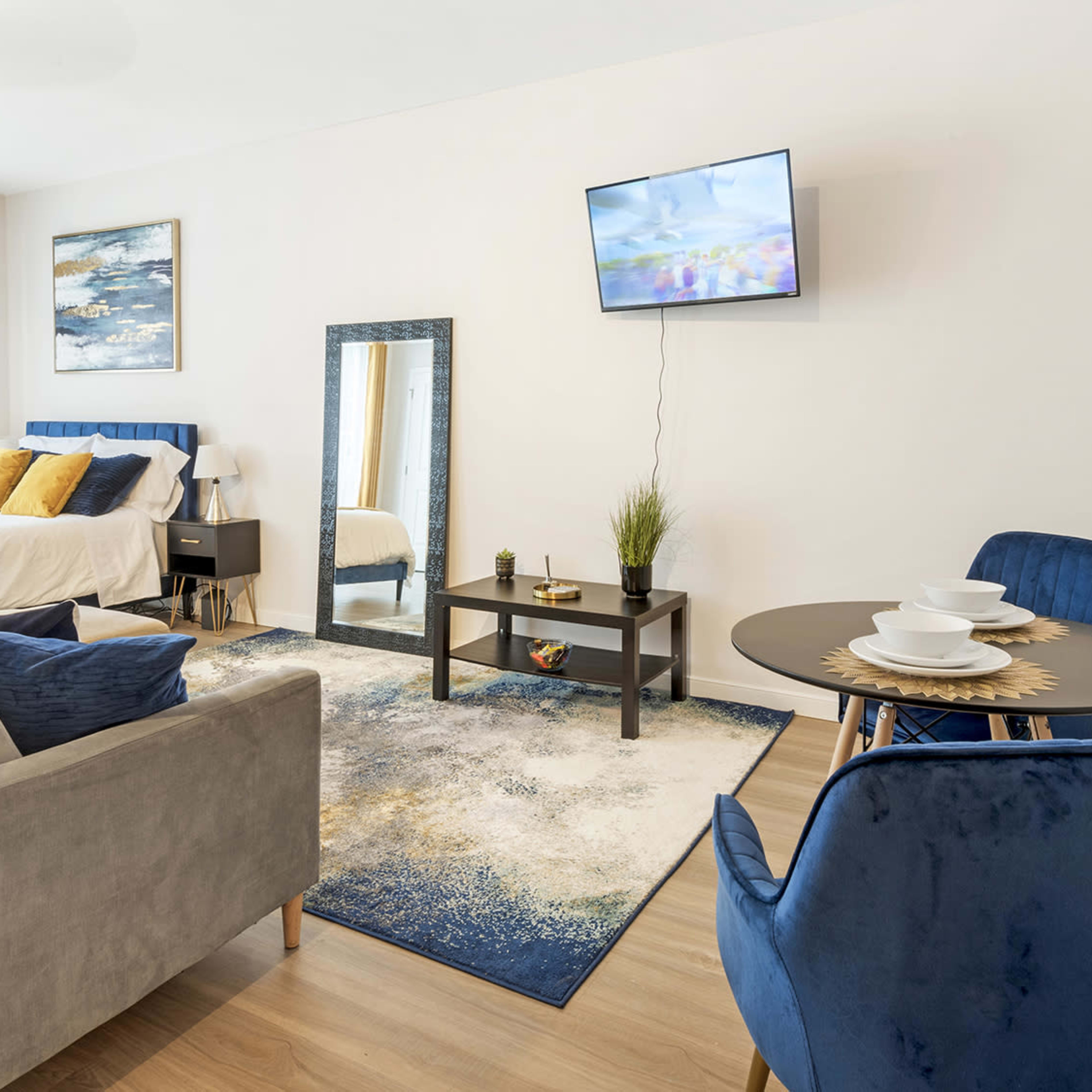 A modern studio apartment with a bed, a television on the wall, a seating area, and a dining table set for two.